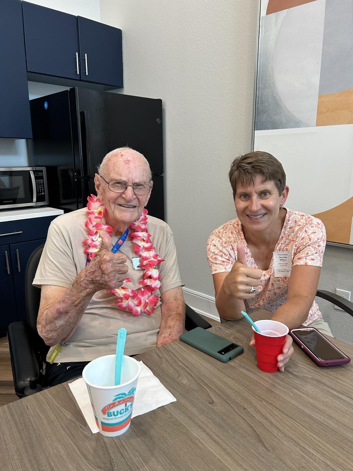 Elderly man wearing a pink lei giving thumbs up sitting at a kitchen table with a staff member also giving thumbs up