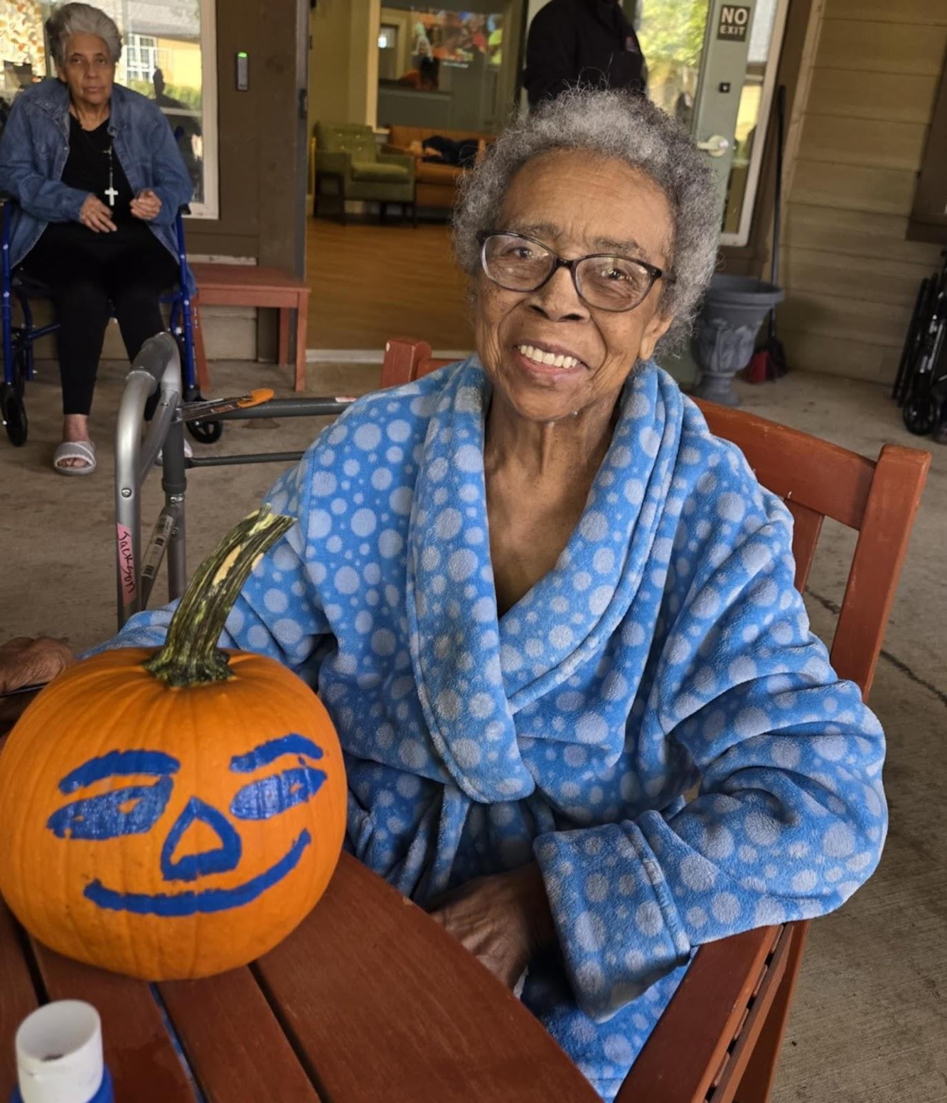 Elderly African American woman in blue patterned robe sitting at table with decorated orange pumpkin, smiling at camera
