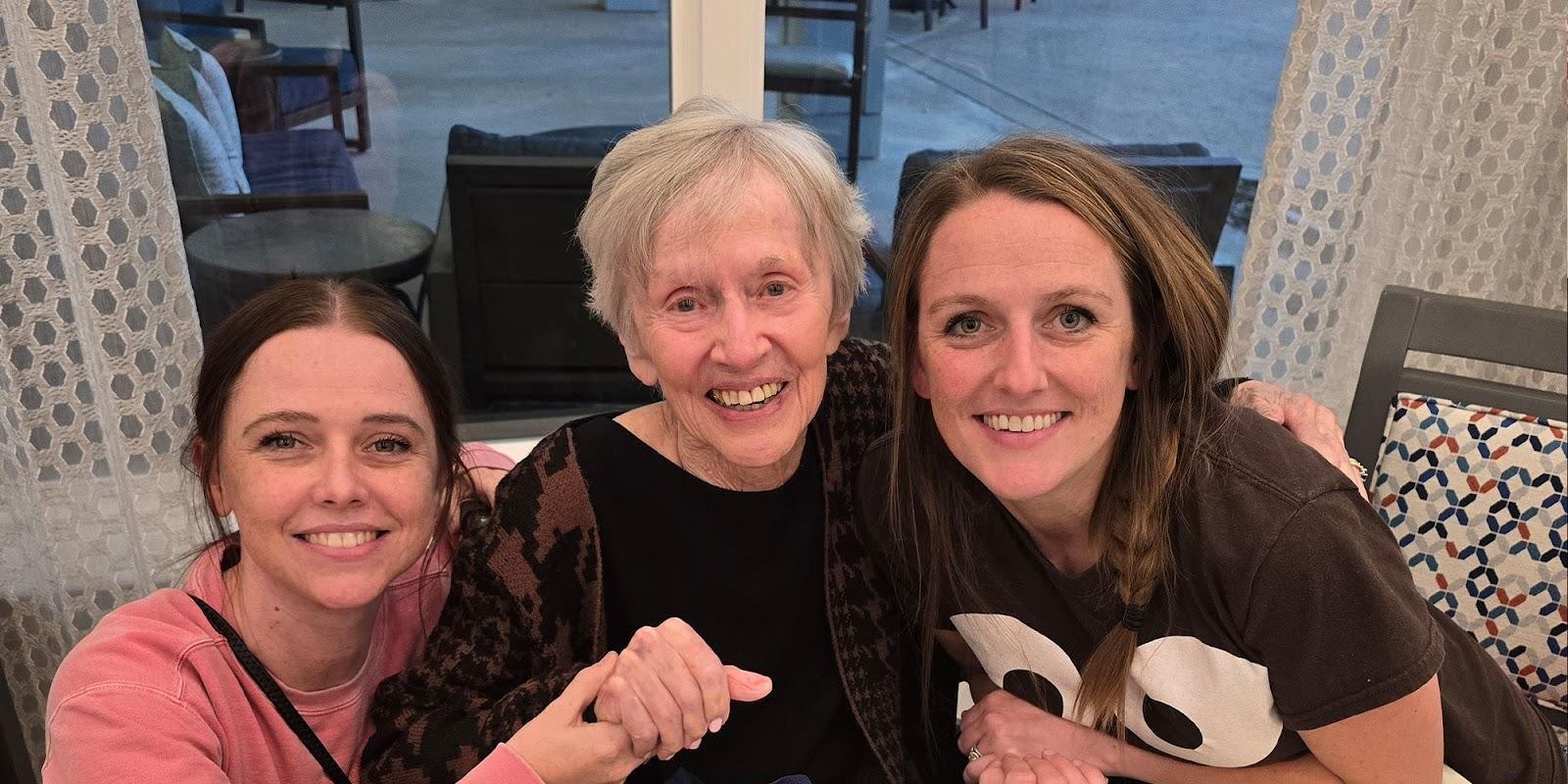 Three women smiling together - elderly woman in center with two younger women on either side in a living room setting