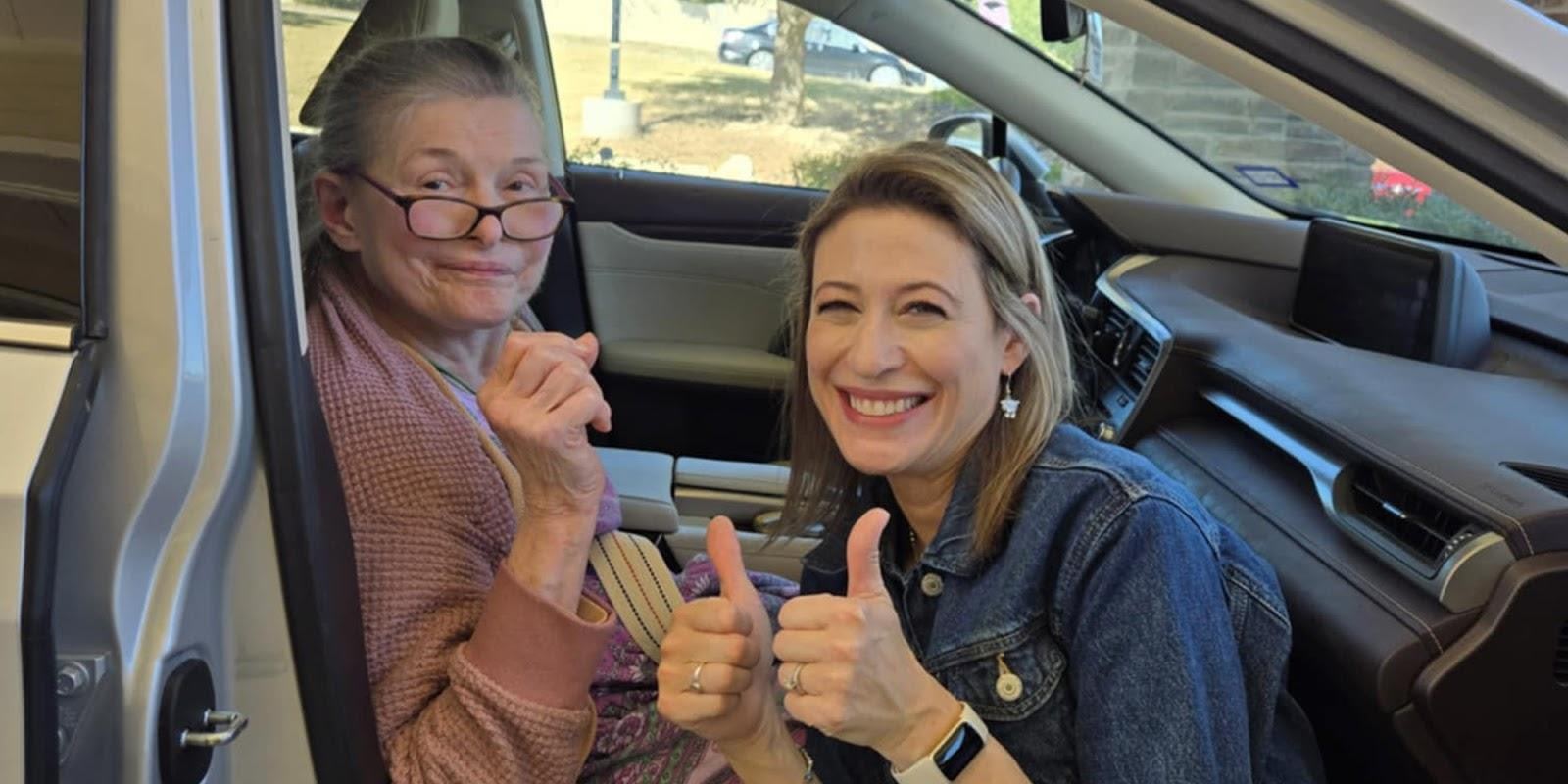 Elderly woman in car giving thumbs up alongside younger woman in driver's seat, both smiling
