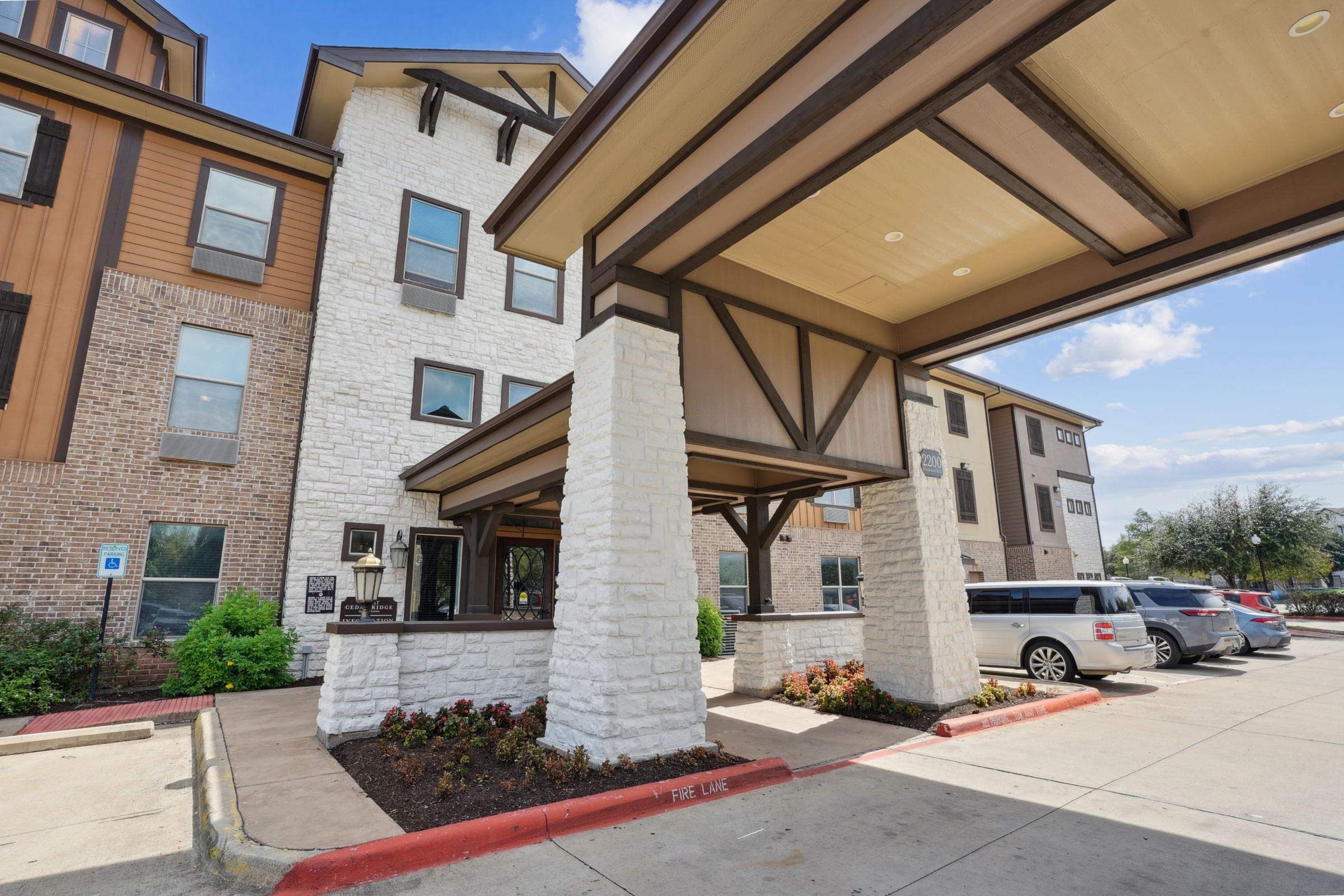 Exterior view of a modern hotel featuring a stone and wood design, with a covered entrance, landscaped flower beds, and parked vehicles in front.