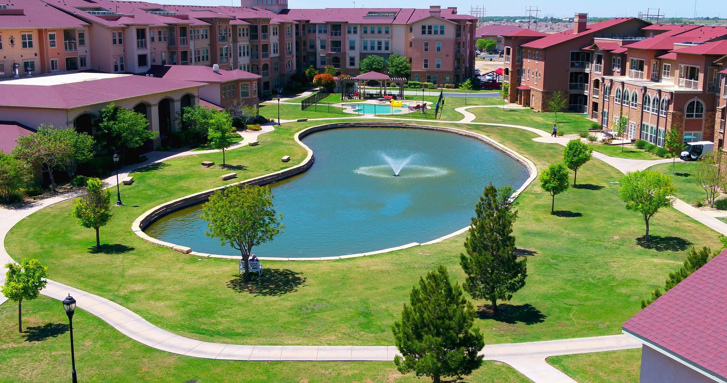 Aerial view of a landscaped courtyard featuring a serene pond with a fountain, surrounded by walking paths and trees, adjacent to modern residential buildings and a swimming pool area.