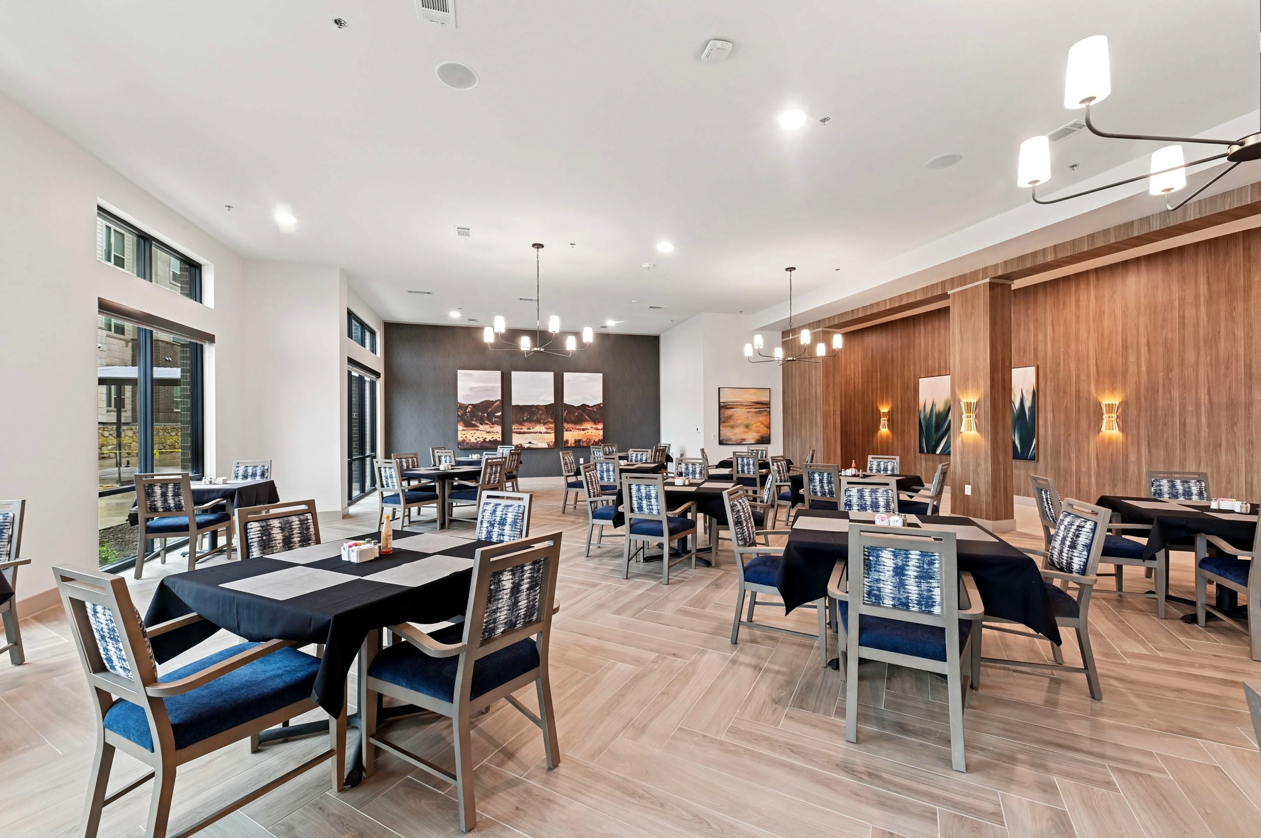 Modern dining area featuring stylish tables with black tablecloths and comfortable chairs, brightened by natural light from large windows. The elegant decor includes wooden accents and artwork, creating a welcoming atmosphere for guests.