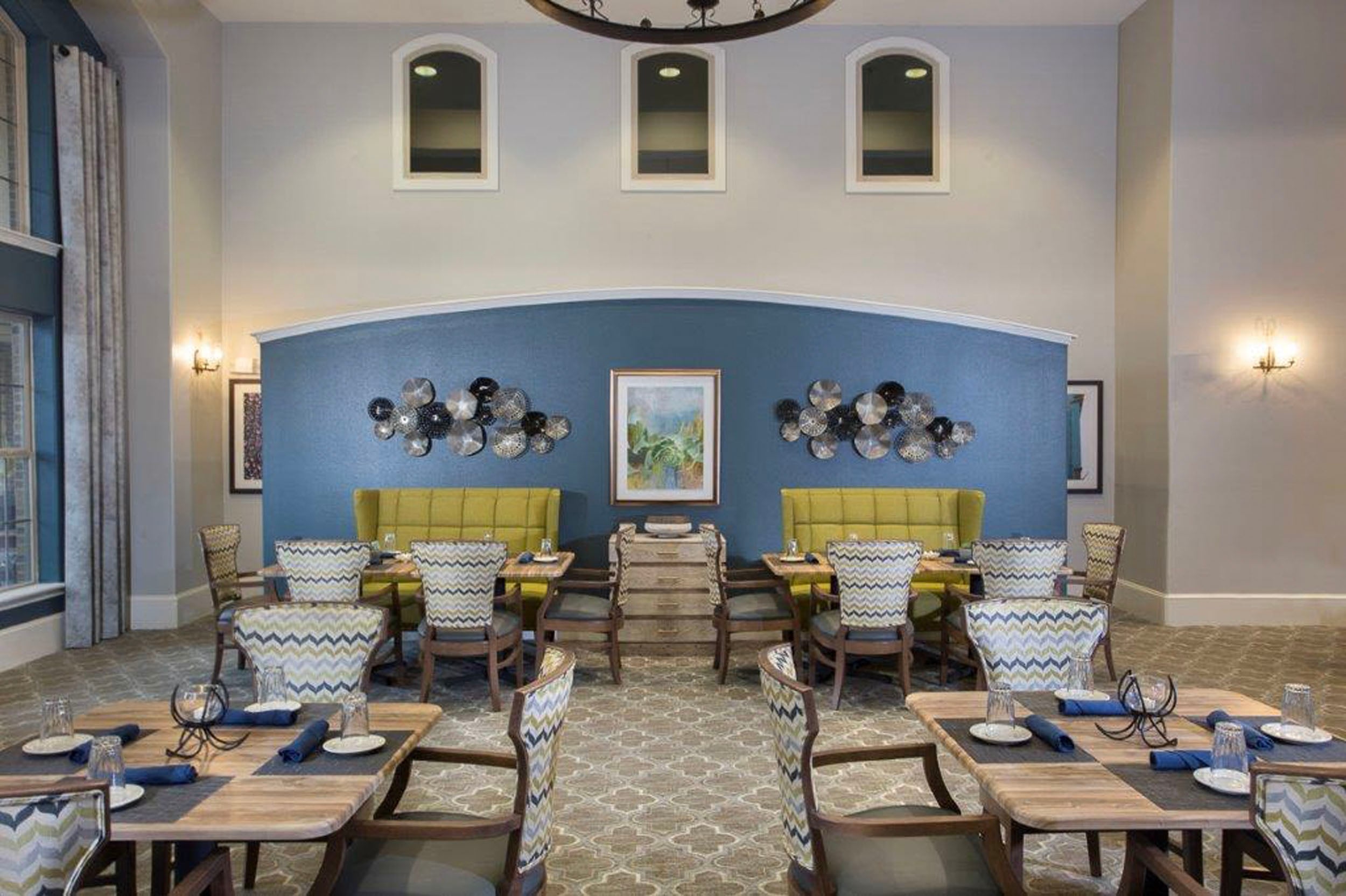 Stylish dining area featuring modern decor with yellow upholstered booths, patterned chairs, and wooden tables, set against a blue accent wall adorned with decorative wall art, complemented by large windows and elegant lighting.