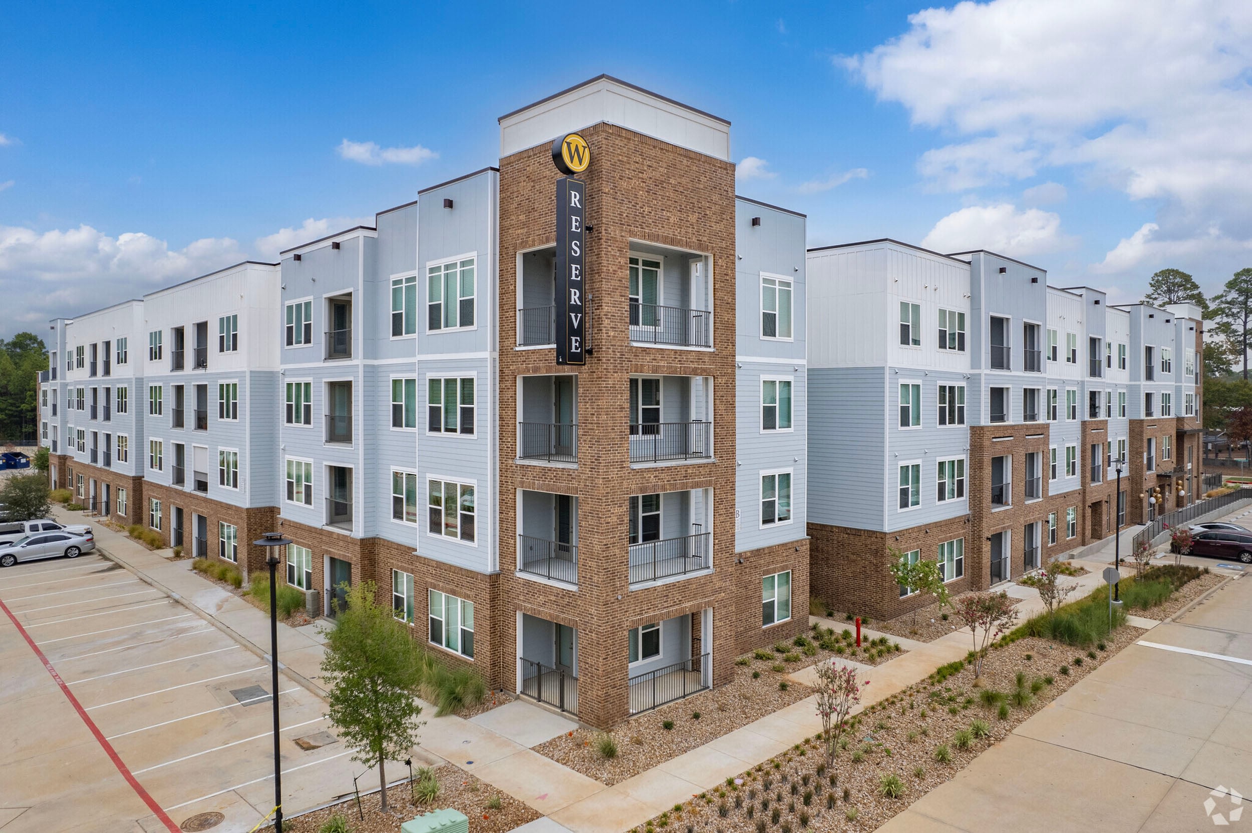Modern apartment complex exterior with brick and siding, featuring balconies and landscaped pathways. Ample parking space in front, showcasing contemporary design and amenities. Ideal for urban living.
