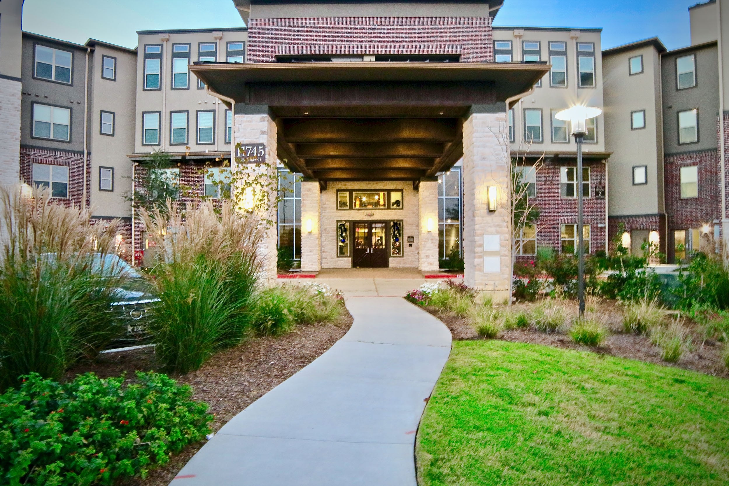 Modern apartment complex entrance with landscaped pathways, greenery, and decorative lighting, showcasing a welcoming atmosphere.