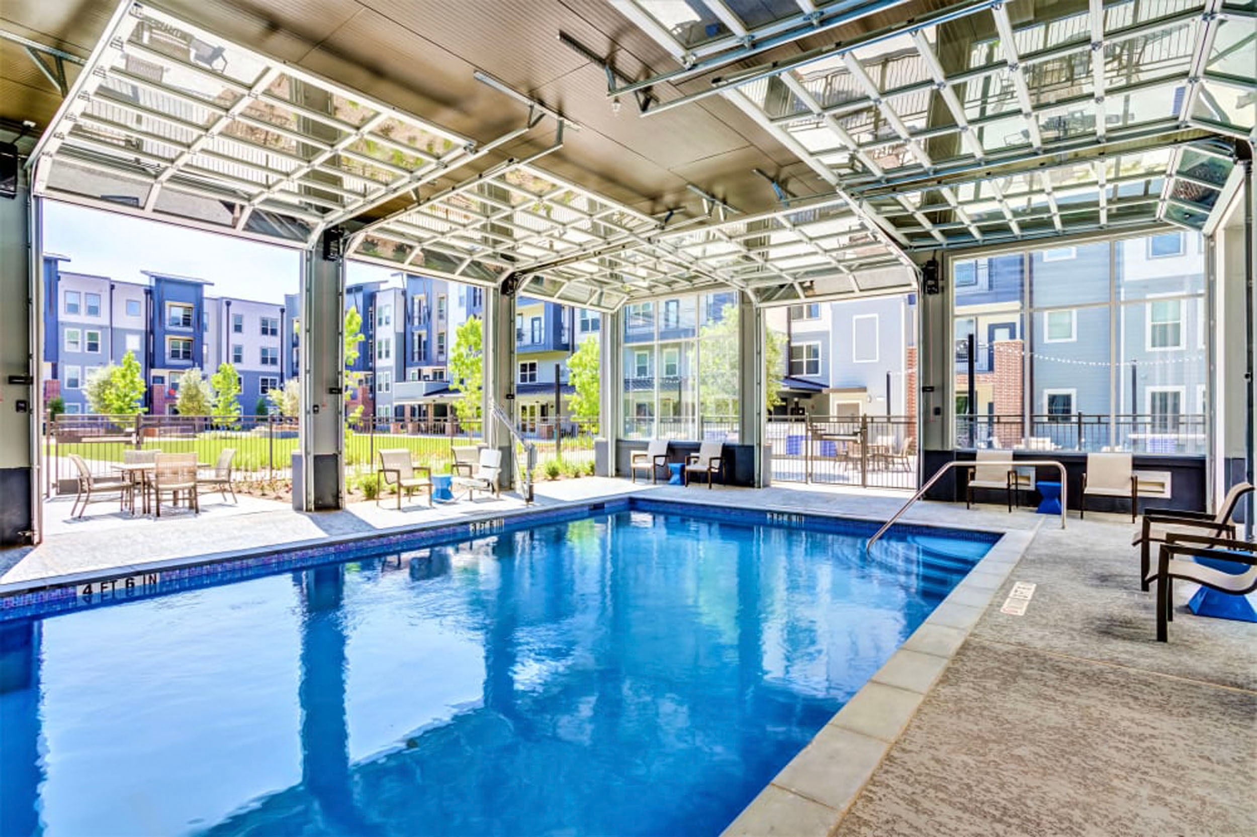 Indoor swimming pool with retractable roof, surrounded by modern apartments and outdoor seating areas, offering a relaxing recreational space.