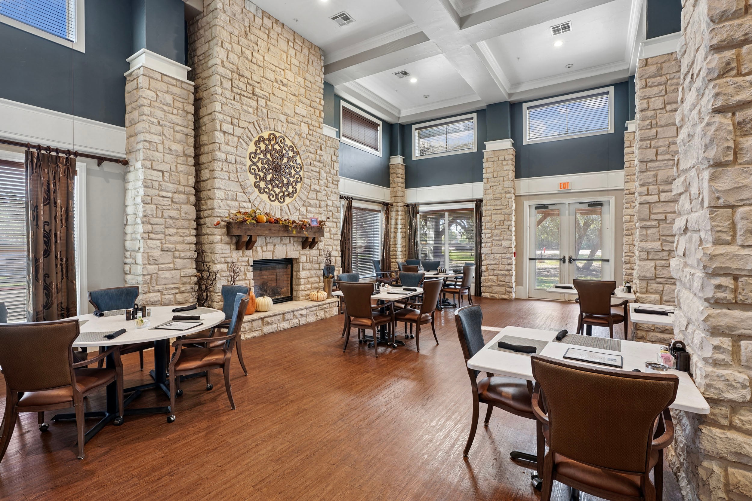 Cozy restaurant interior featuring a stone fireplace, wooden flooring, and elegant dining tables with chairs. The space is well-lit with large windows and decorative elements, creating a warm and inviting atmosphere for guests.