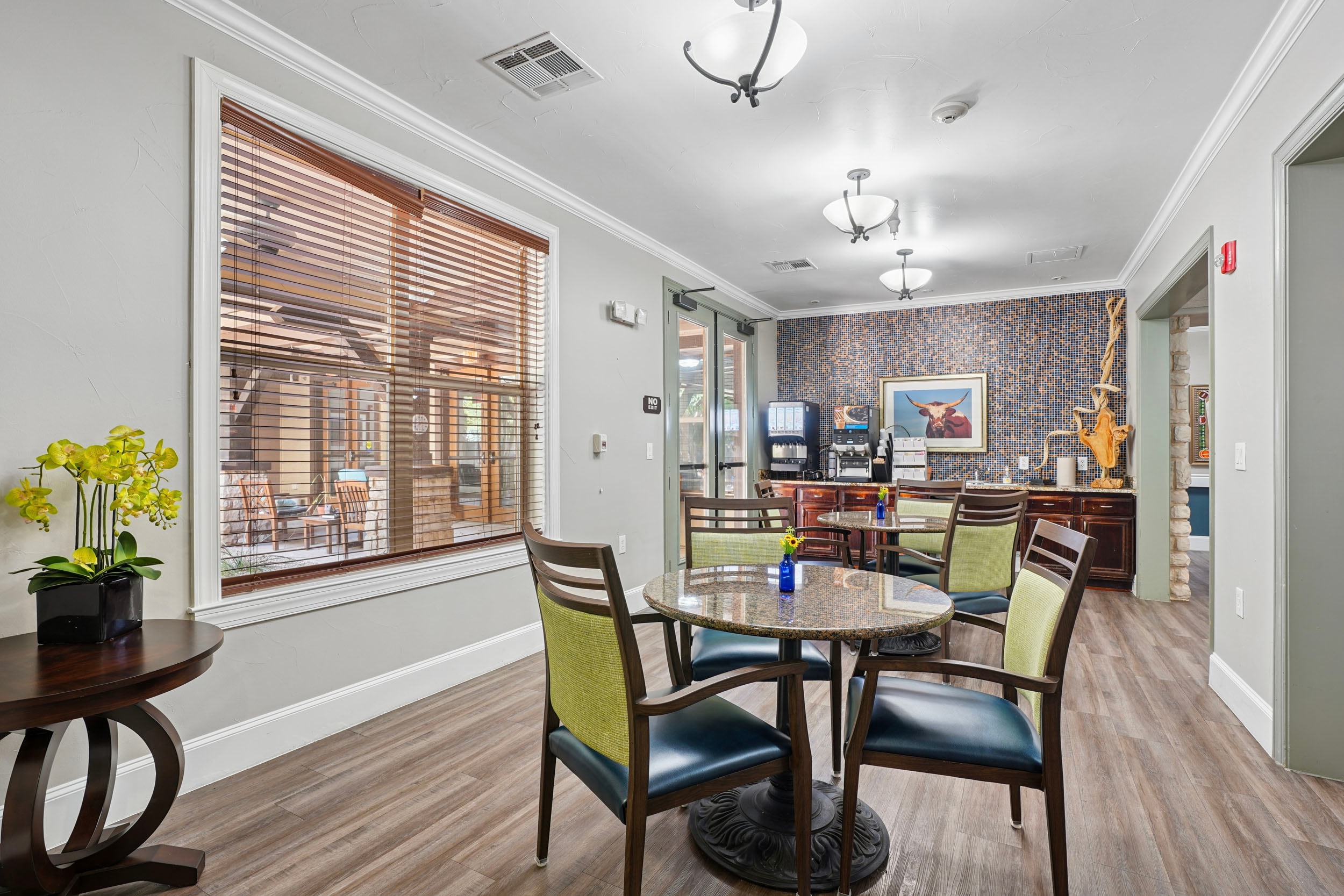 Bright and inviting common area featuring a round table with colorful chairs, a decorative wall with art, and a large window with blinds. A coffee station is visible in the background, complemented by a potted orchid on a side table. The space is designed for relaxation and socializing.