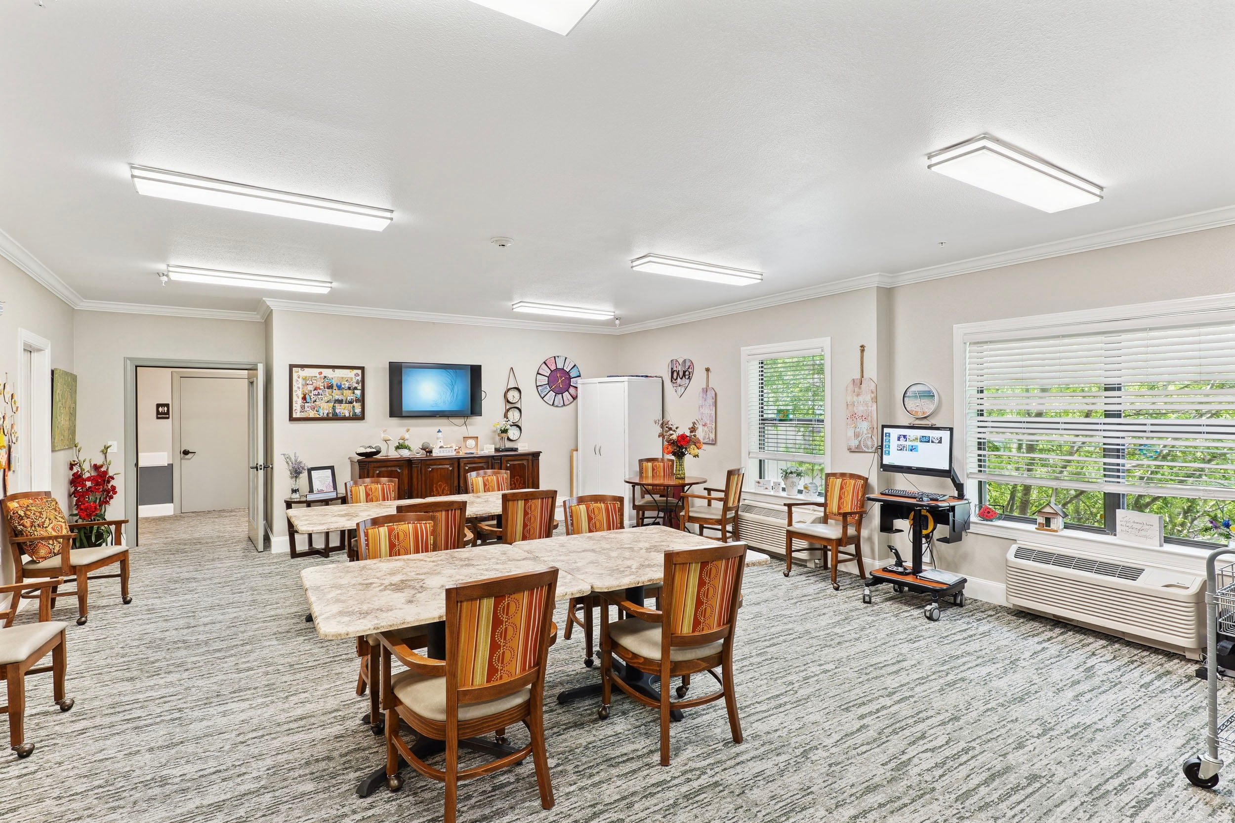 Bright and inviting communal activity room featuring wooden tables and chairs, a large flat-screen TV, decorative wall art, and windows providing natural light. The space is designed for group activities and social interaction, with a cozy atmosphere enhanced by plants and decorative elements.