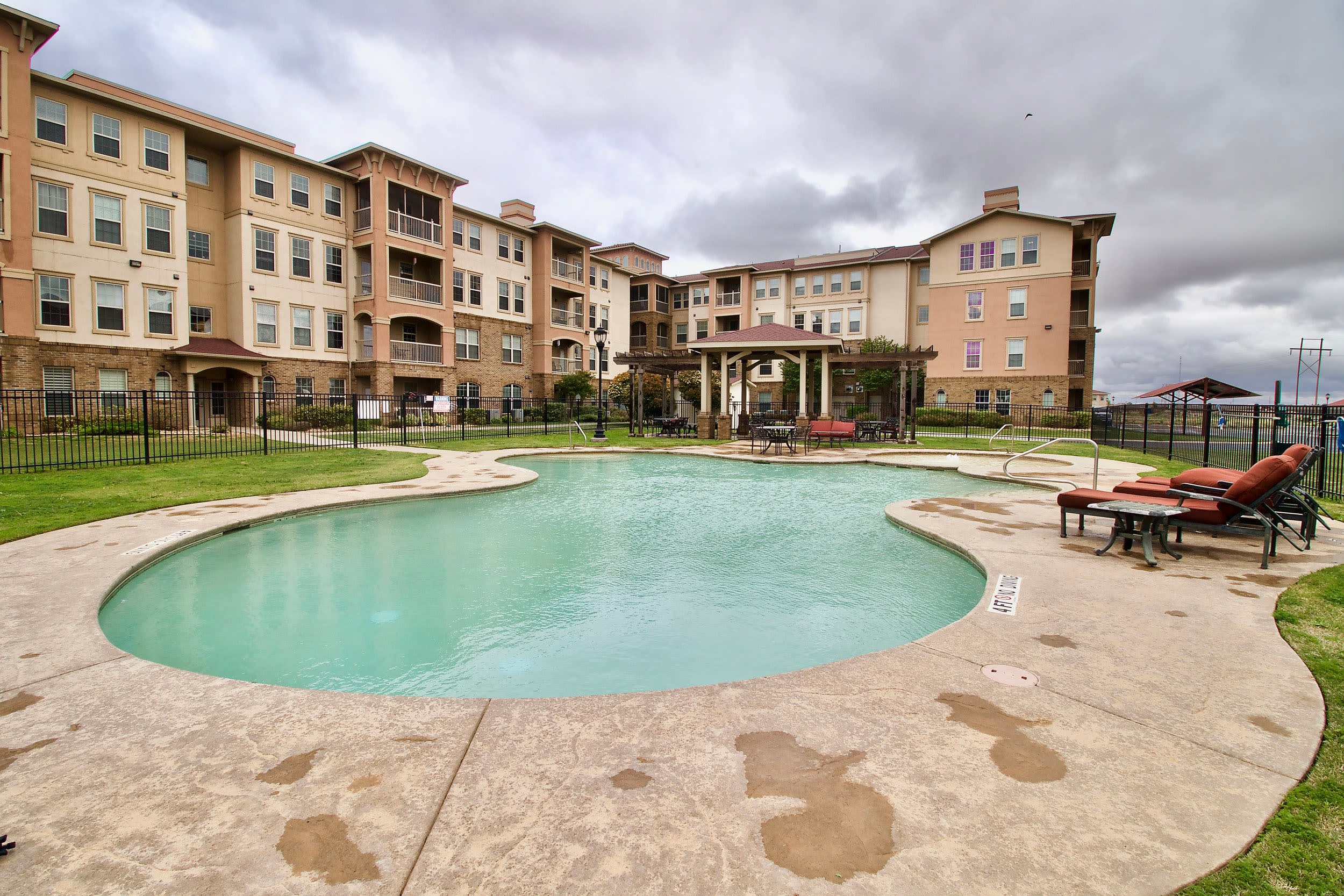Apartment complex with a swimming pool and lounge area, featuring a pergola and seating, surrounded by green lawn and a fence, under a cloudy sky.