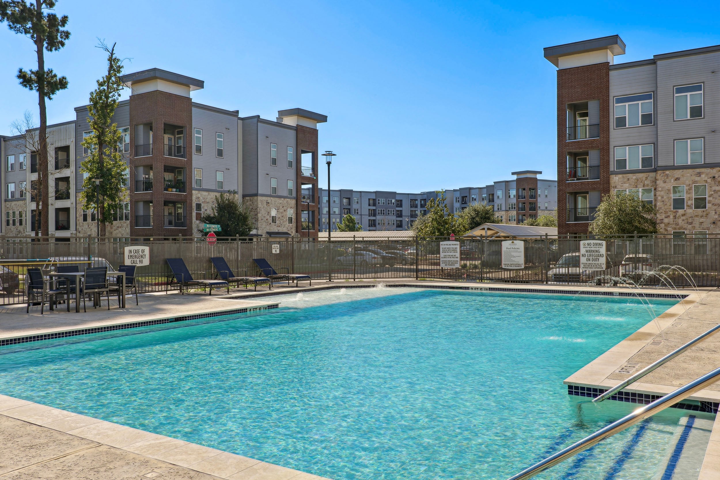 Modern apartment complex with a sparkling swimming pool, surrounded by sun loungers and a secure fence, offering a relaxing outdoor space in a vibrant community.