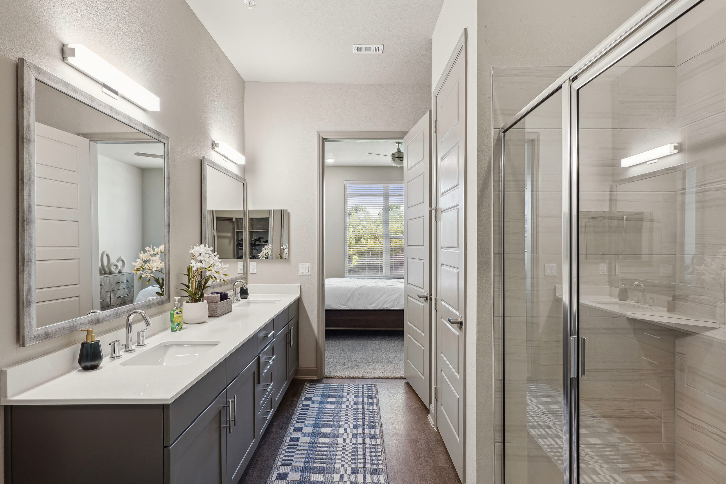 Modern bathroom interior featuring dual sinks, large mirrors, and a glass shower. The space is well-lit with contemporary fixtures, neutral wall colors, and decorative elements like a potted plant. A doorway leads to a bedroom with natural light filtering through window blinds.