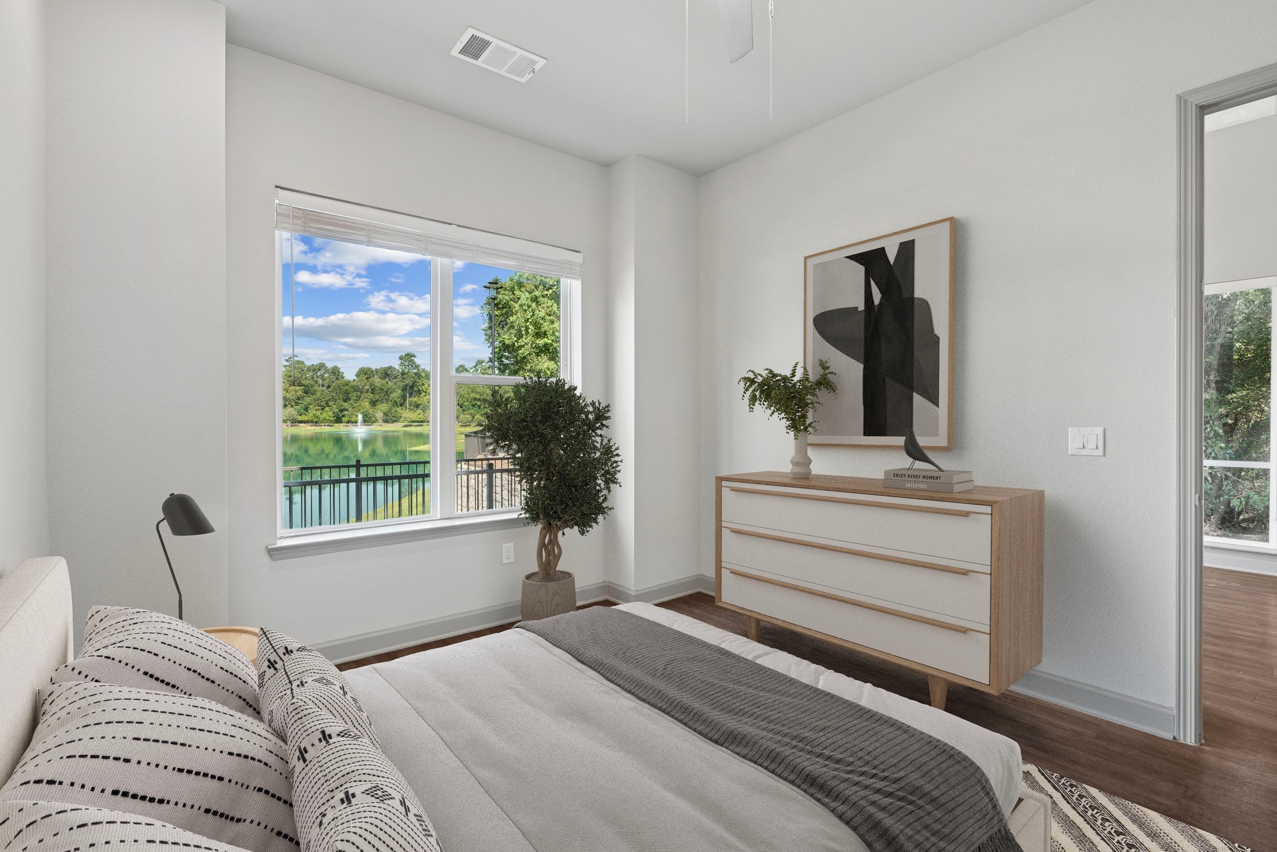 Modern bedroom with a large window showcasing a serene view of a lake and greenery. The room features a cozy bed with patterned pillows, a stylish dresser, and decorative plants, creating a tranquil and inviting atmosphere.