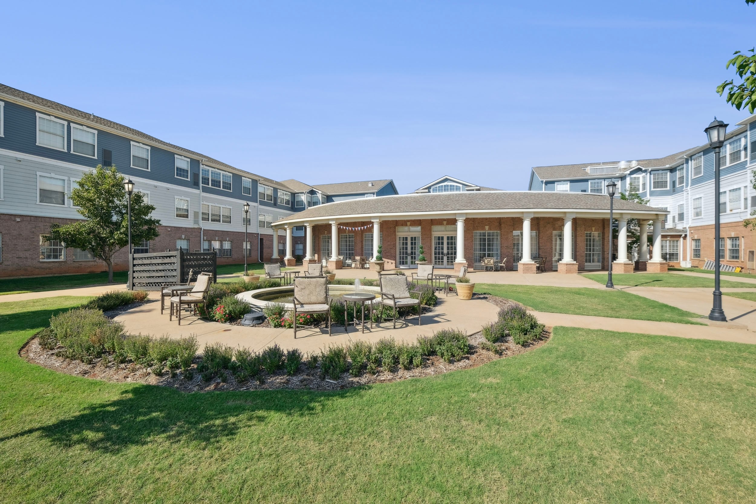 Courtyard of a modern residential community featuring a circular seating area, landscaped gardens, and a gazebo-like structure, surrounded by multi-story buildings under a clear blue sky.