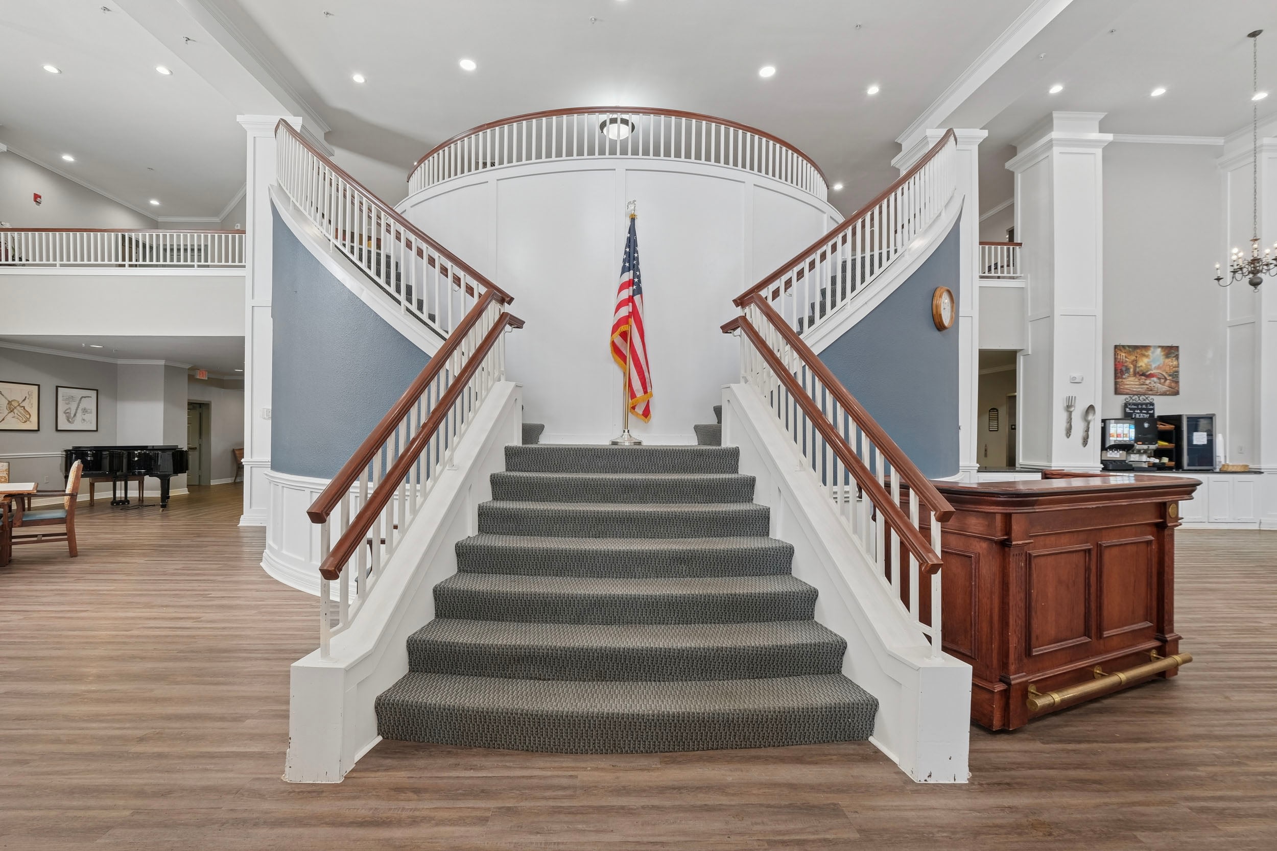 Elegant staircase with a carpeted runner leading to a second-floor landing, featuring a decorative railing and an American flag displayed at the center. The background includes a spacious lobby area with wooden flooring, a piano, and a reception desk, showcasing a welcoming atmosphere.
