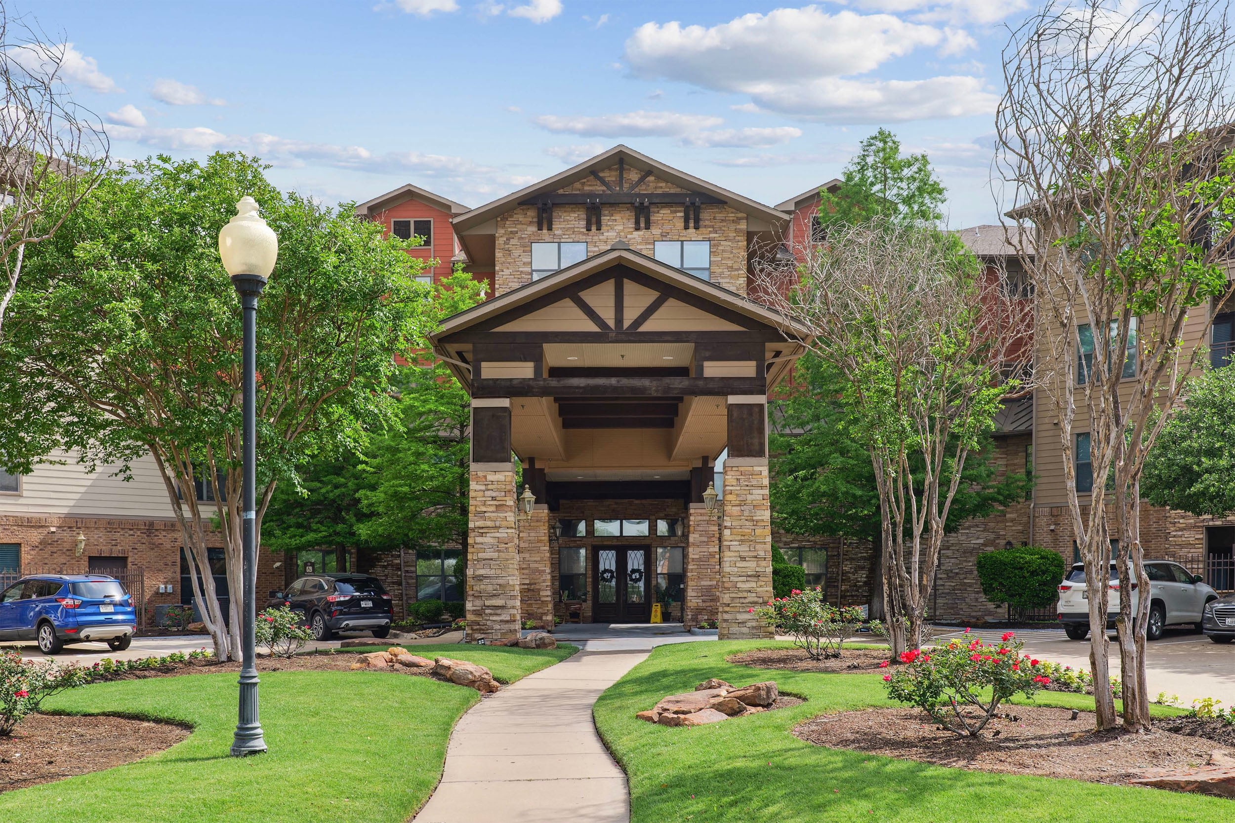 Entrance of a modern apartment complex featuring stone architecture, landscaped gardens, and a paved walkway, surrounded by lush greenery and parked cars. Ideal for potential renters or visitors.