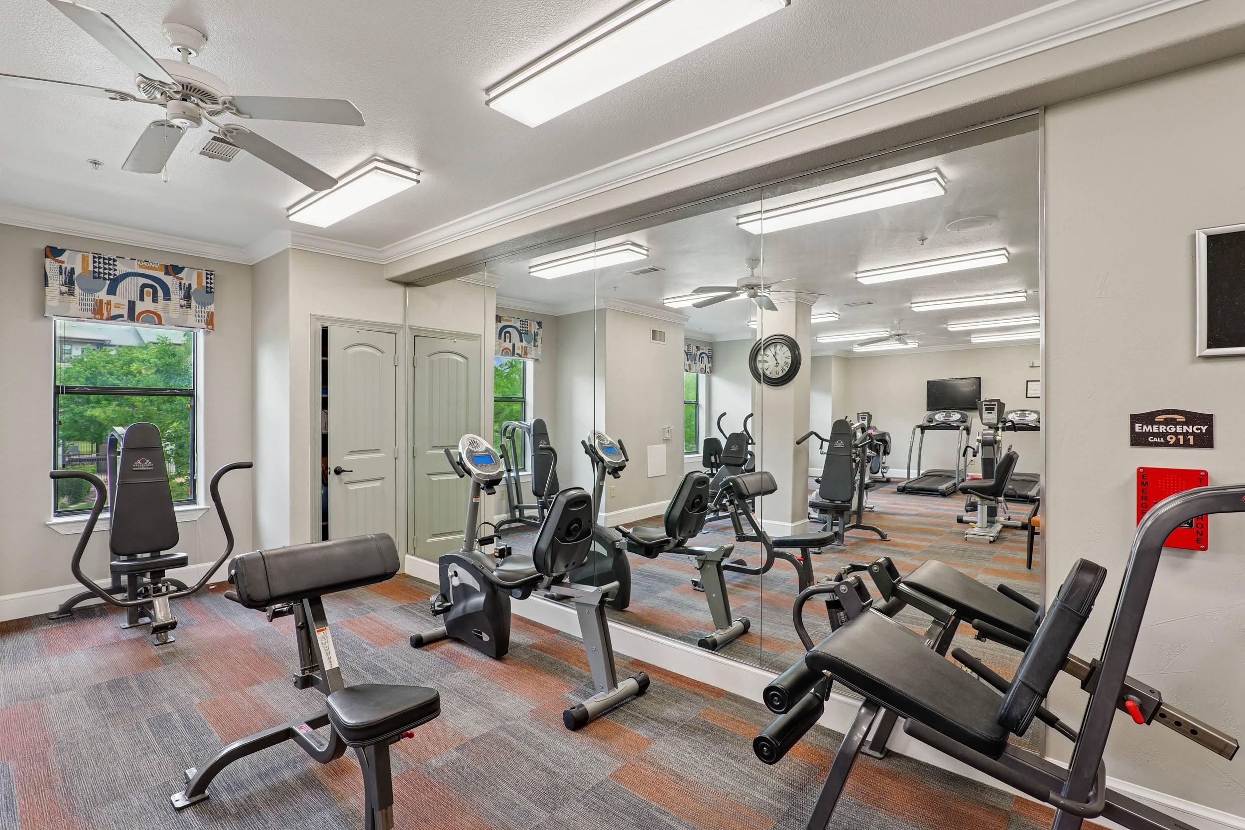 Modern fitness center with exercise equipment, including stationary bikes and weight benches, surrounded by mirrors and bright lighting, featuring large windows for natural light and a ceiling fan for ventilation.