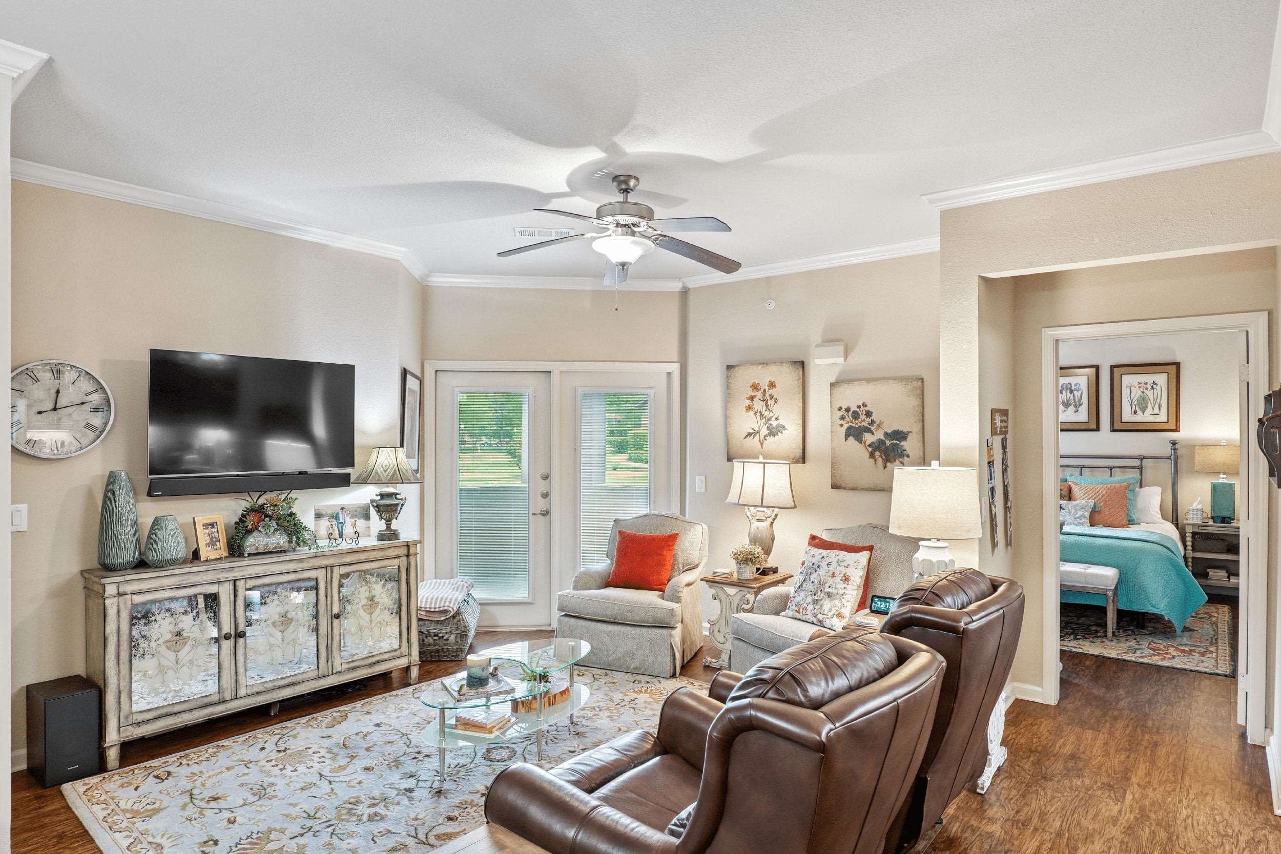 Cozy living room featuring a modern design with a flat-screen TV mounted on the wall, stylish furniture including leather recliners, and decorative accents. The space is illuminated by natural light from sliding glass doors and lamps, complemented by a patterned area rug and artwork. A glimpse of a bedroom is visible through an adjoining doorway.