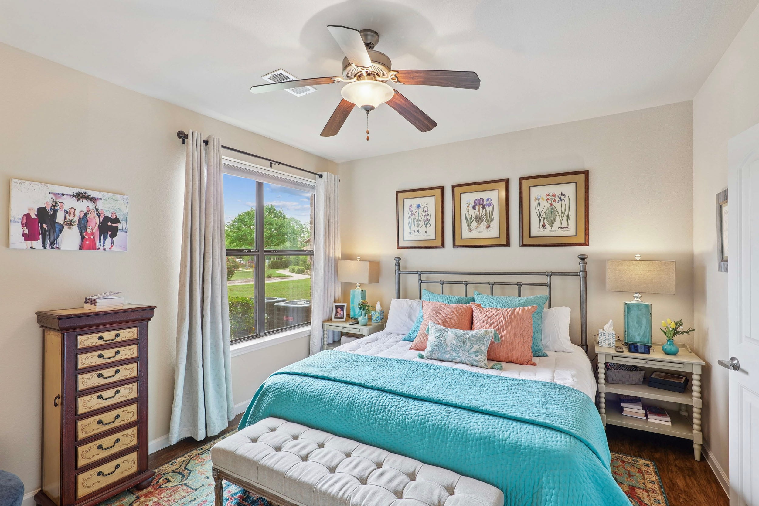 Cozy bedroom featuring a stylish metal bed frame adorned with turquoise and coral bedding, a tufted bench at the foot, and decorative pillows. The room includes a wooden dresser, bedside table with a lamp, and framed botanical art on the walls. Natural light floods in through a large window, enhancing the inviting atmosphere.