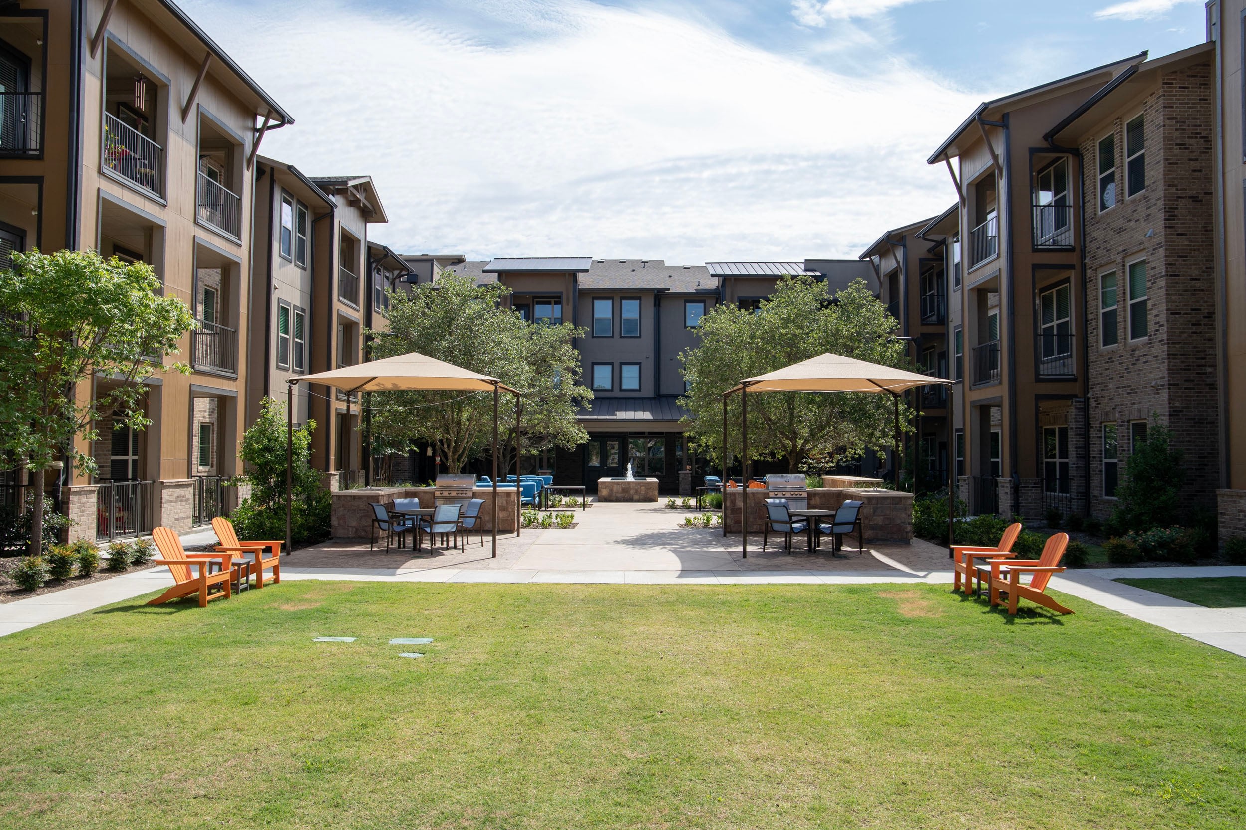 Modern apartment complex courtyard featuring green grass, shaded seating areas, and landscaped trees, ideal for relaxation and outdoor gatherings.