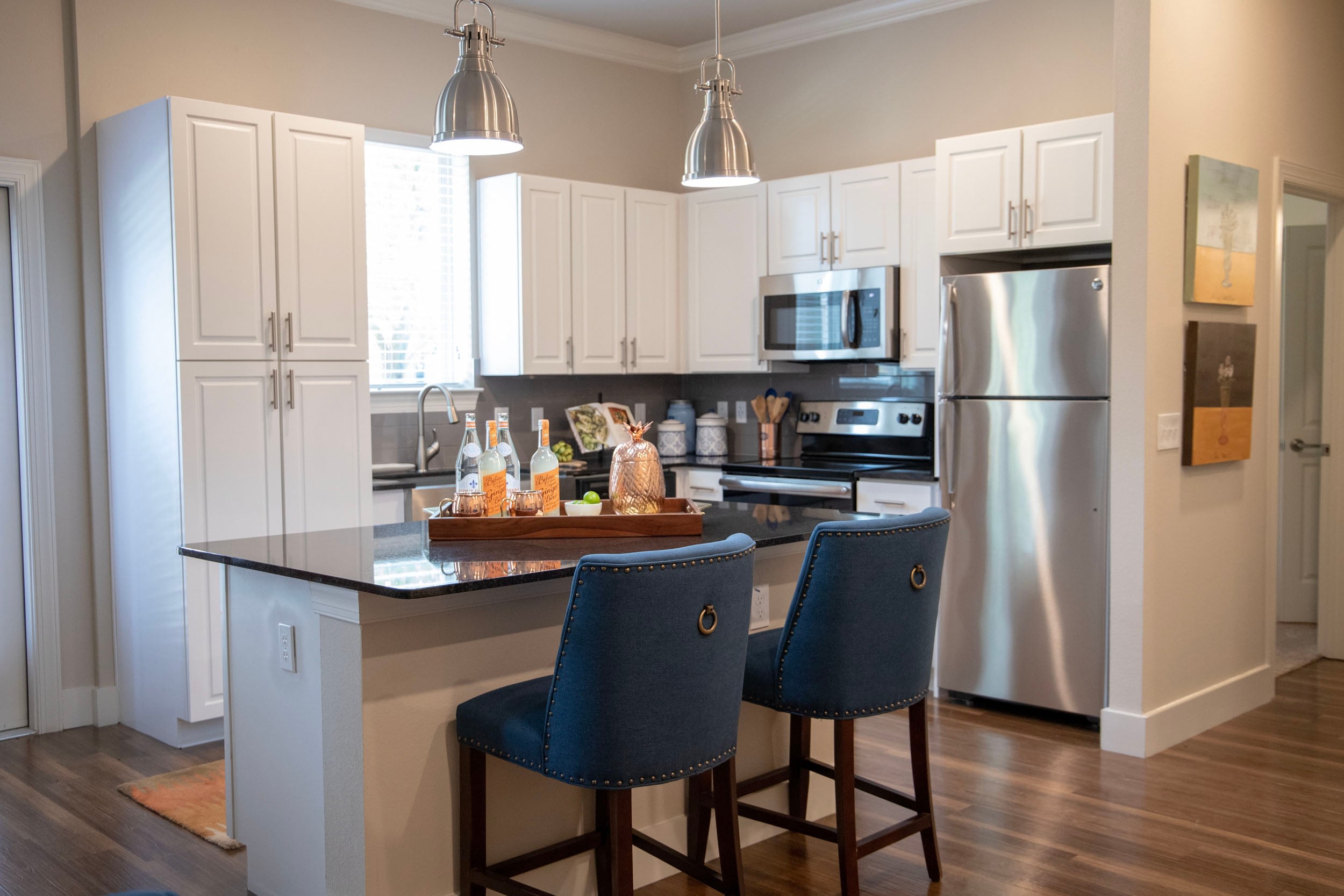 Modern kitchen interior featuring white cabinetry, stainless steel appliances, and a dark granite countertop. The space includes a kitchen island with two blue upholstered bar stools and a wooden tray displaying drinks and decor. Natural light streams in through the window, enhancing the inviting atmosphere.