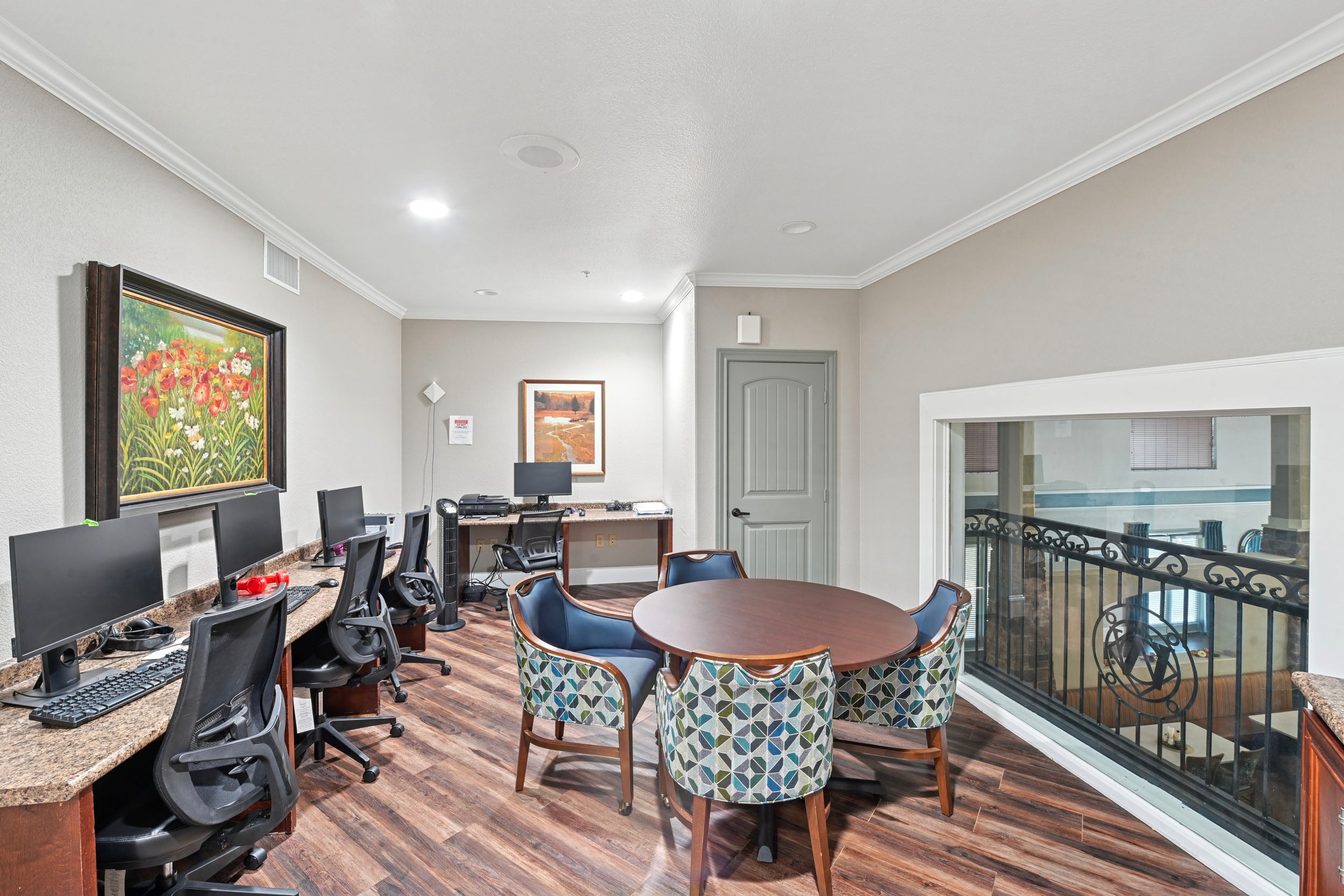 Modern office space featuring multiple computer stations with ergonomic chairs, a round wooden table surrounded by patterned chairs, and decorative artwork on the walls. The room is well-lit with natural light coming from a large window, creating an inviting atmosphere for work and collaboration.
