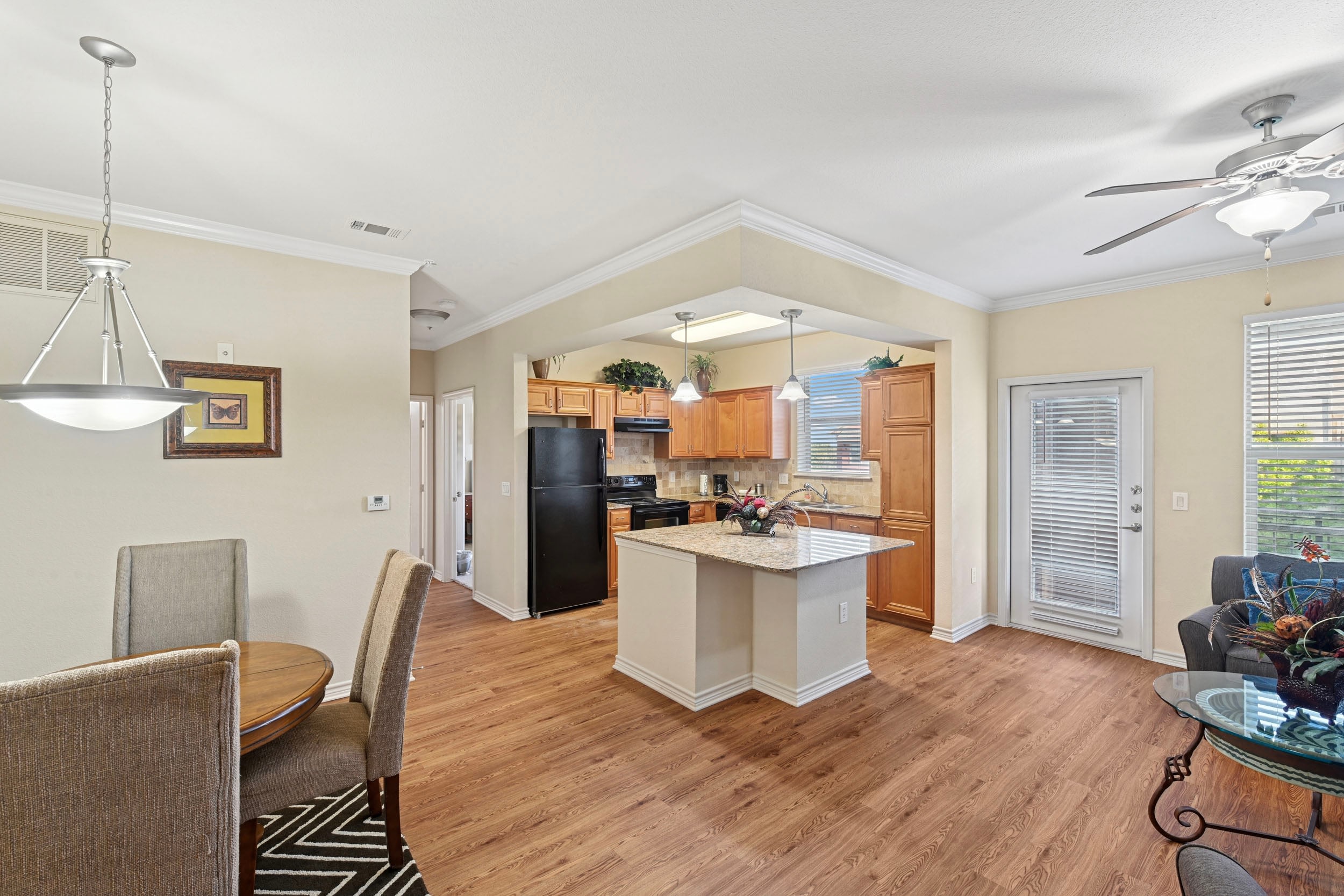 Modern kitchen and dining area with hardwood floors, featuring a round wooden table with upholstered chairs, a granite countertop island, and stainless steel appliances. The space is well-lit with natural light from windows and stylish pendant lighting.