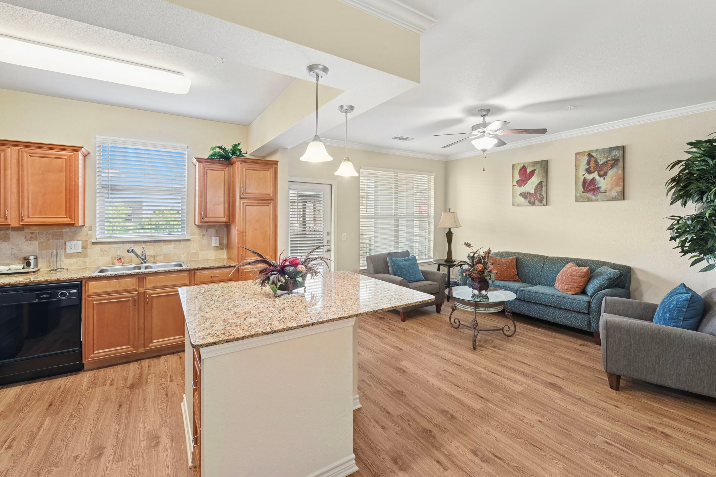 Modern kitchen and living room interior featuring wooden cabinetry, granite countertops, and a cozy seating area with decorative cushions. The space is well-lit with natural light from large windows, complemented by stylish decor including artwork and potted plants.
