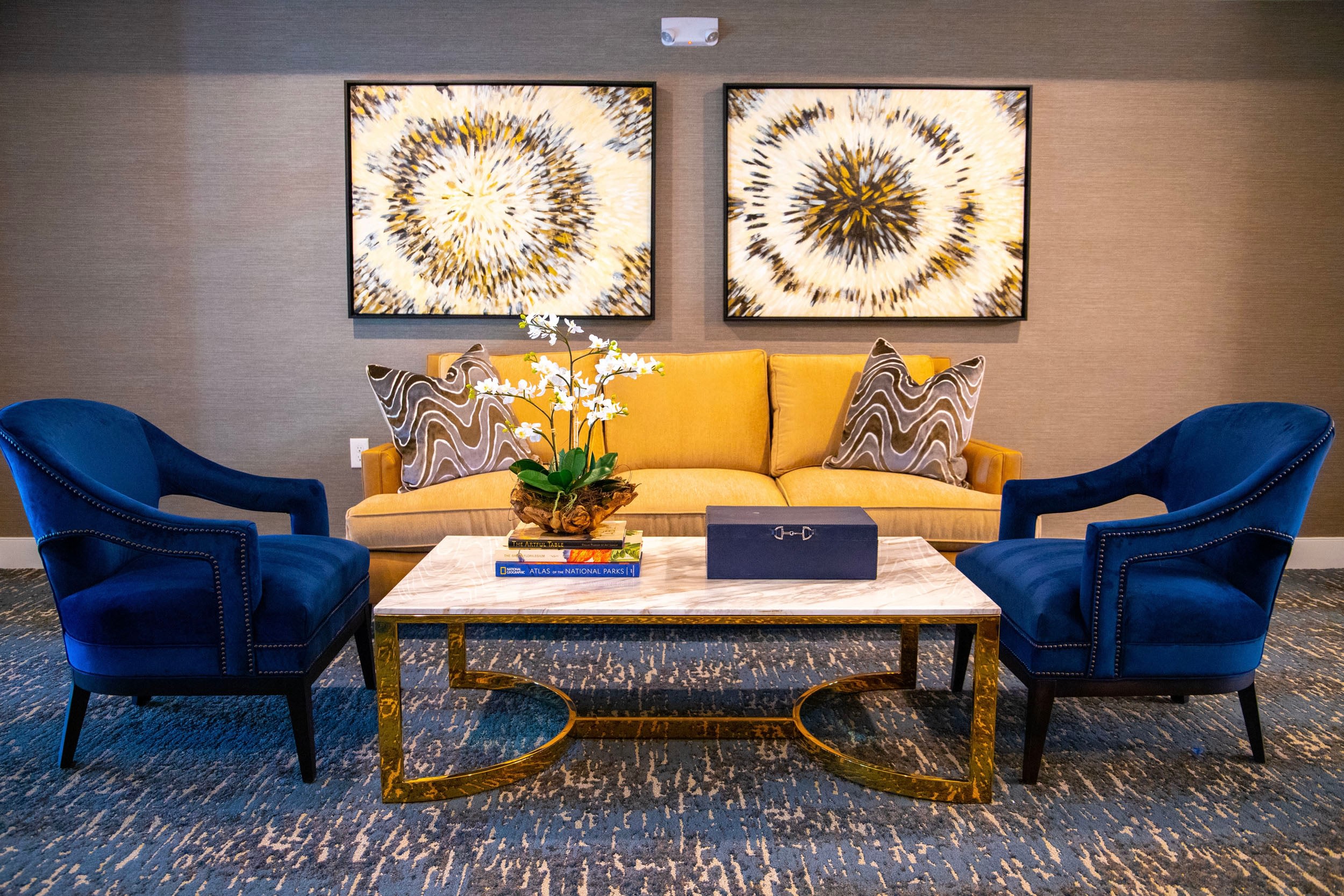 Stylish living room setup featuring a yellow sofa with decorative pillows, two blue accent chairs, and a marble coffee table adorned with a potted orchid and books, complemented by abstract wall art.
