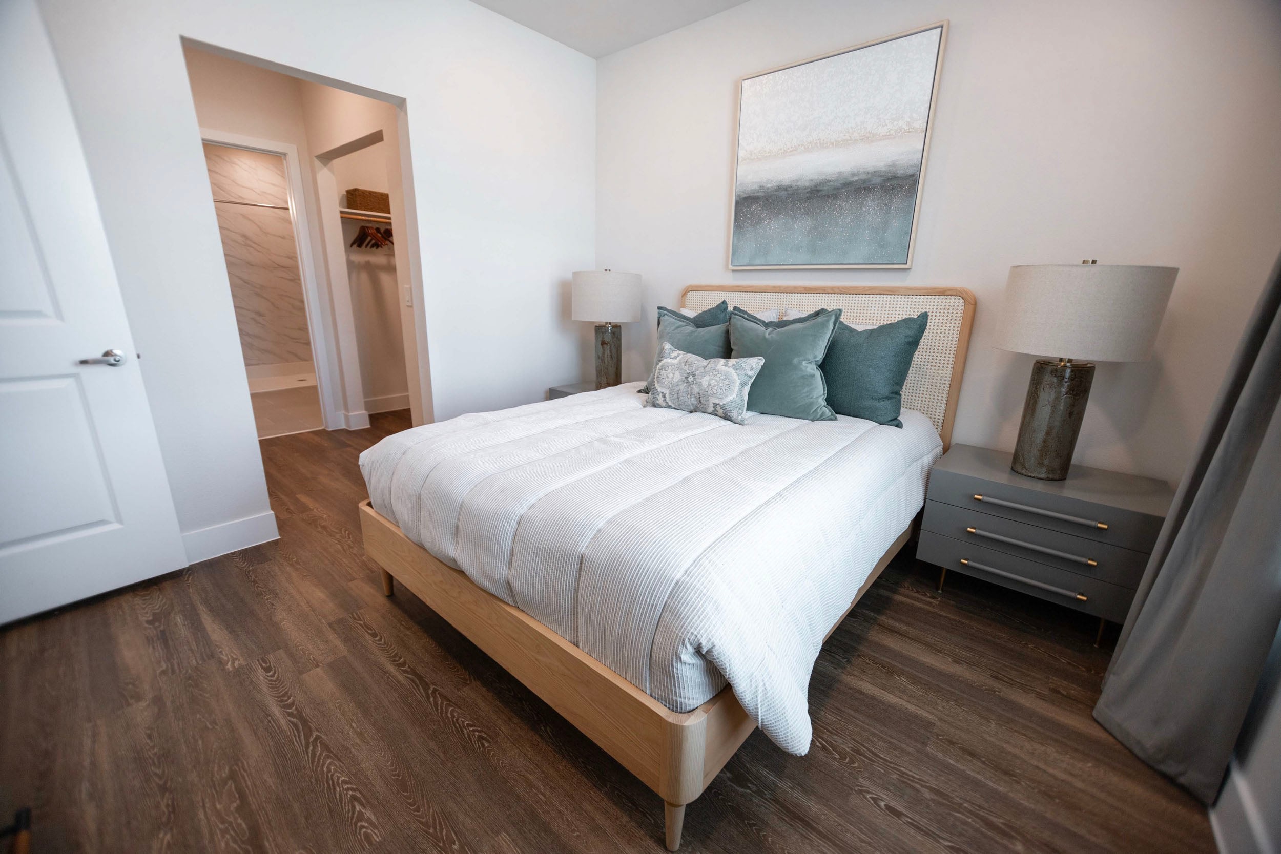 Modern bedroom featuring a comfortable bed with decorative pillows, a stylish nightstand, and a wall art piece. The room has a warm wooden floor and a doorway leading to a bathroom area, creating a cozy and inviting atmosphere.