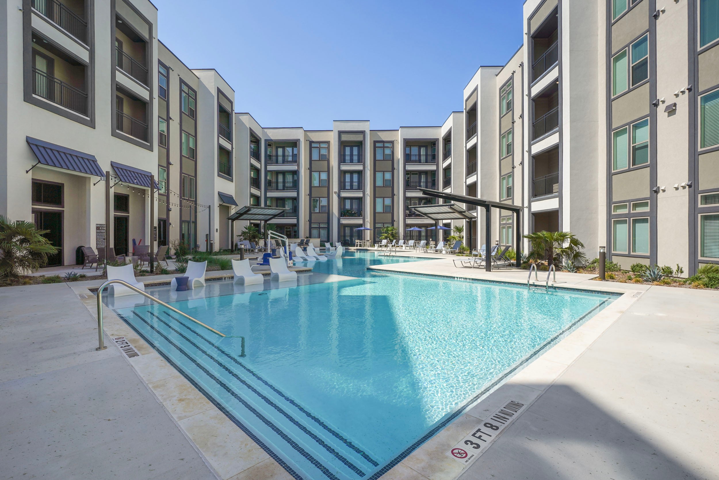 Modern apartment complex with a sparkling swimming pool surrounded by lounge chairs and palm trees, featuring spacious balconies and large windows in a sunny outdoor setting.