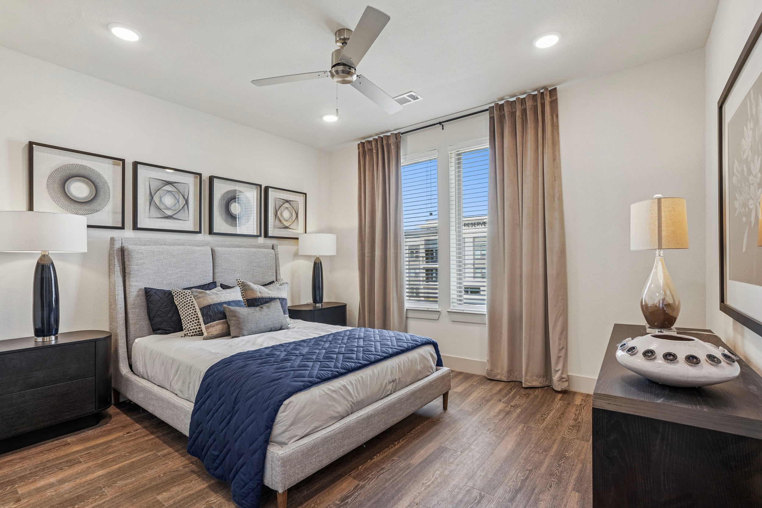 Modern bedroom featuring a light gray upholstered bed with navy blue bedding, decorative pillows, and stylish wall art. Includes dark wood nightstands with lamps, large windows with blinds, and elegant curtains, creating a bright and inviting atmosphere.
