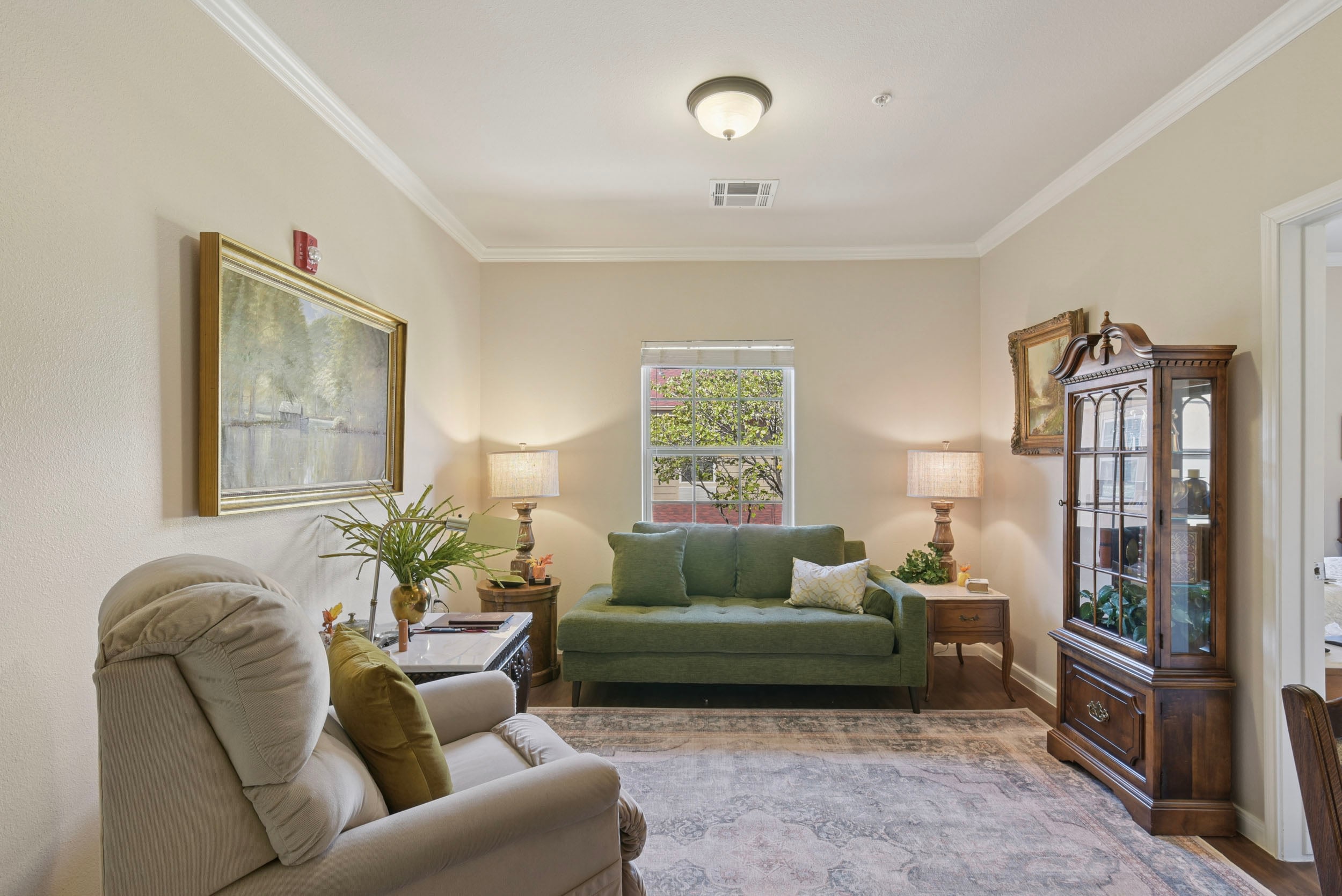 Cozy living room featuring a green sofa, beige armchair, decorative wooden furniture, and a large window with natural light. The walls are adorned with artwork and the room includes houseplants, creating a welcoming atmosphere.