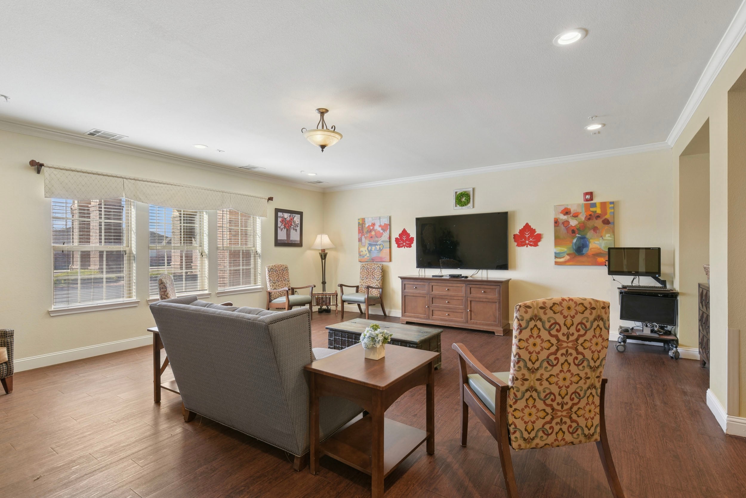 Cozy living room featuring comfortable seating, a wooden coffee table, and a large TV. Natural light streams through multiple windows with blinds, enhancing the inviting atmosphere. Decorated with vibrant artwork and a lamp for additional lighting, this space is perfect for relaxation or social gatherings.