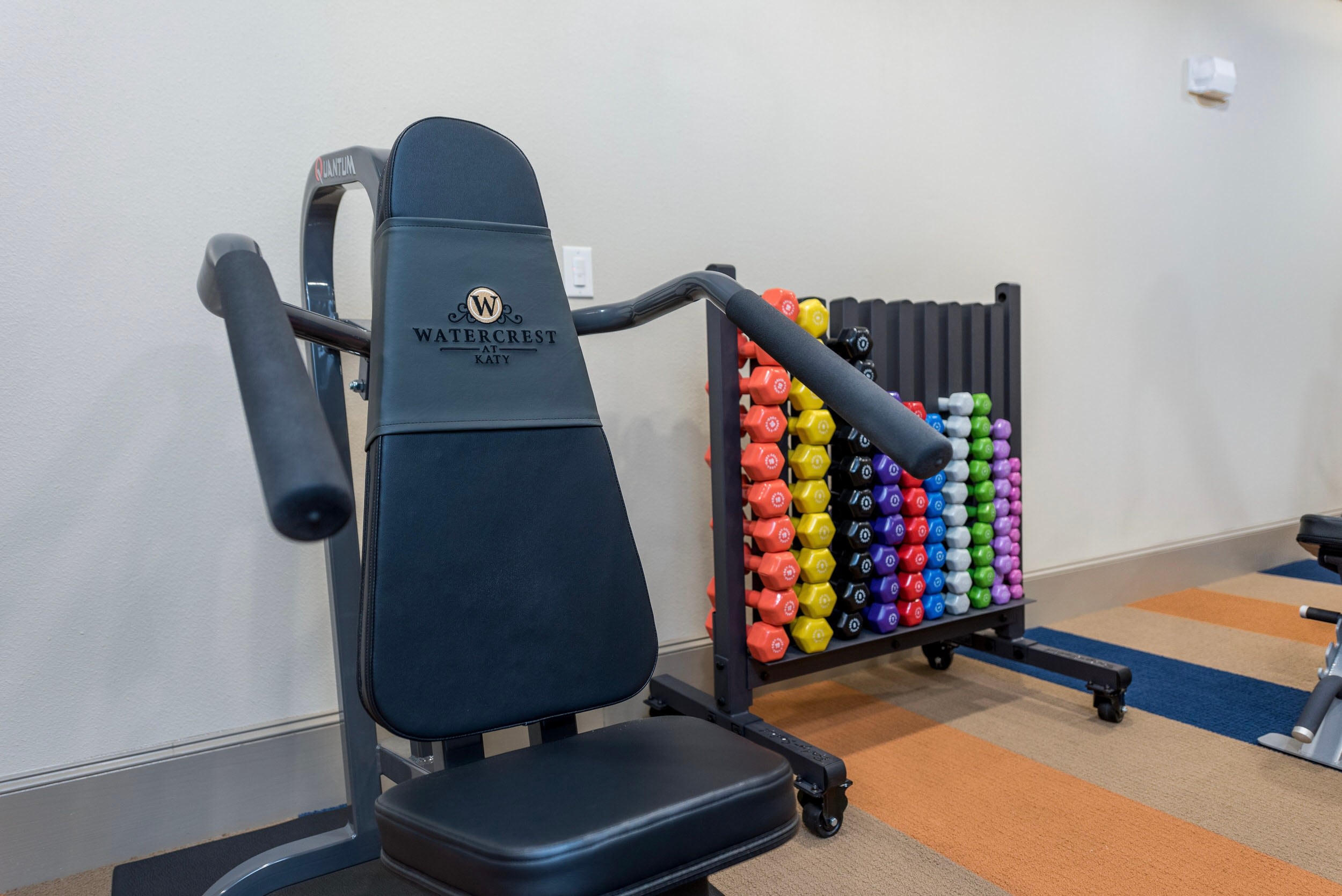 Gym equipment at Watercrest at Katy featuring a weight bench and a colorful rack of dumbbells in a fitness center. The space is designed for strength training and fitness activities.