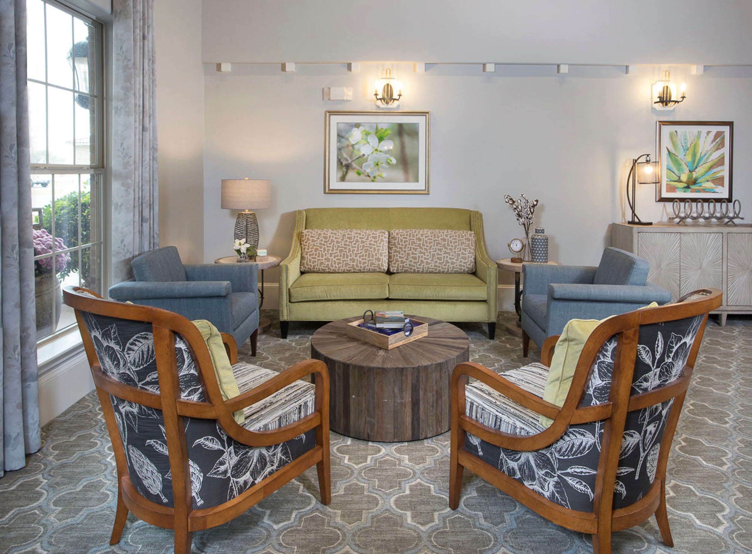 Cozy living room featuring a green sofa with patterned cushions, two blue armchairs, and two wooden chairs with floral upholstery. A round wooden coffee table sits in the center, complemented by decorative accents and wall art. Natural light filters through large windows, enhancing the inviting atmosphere.
