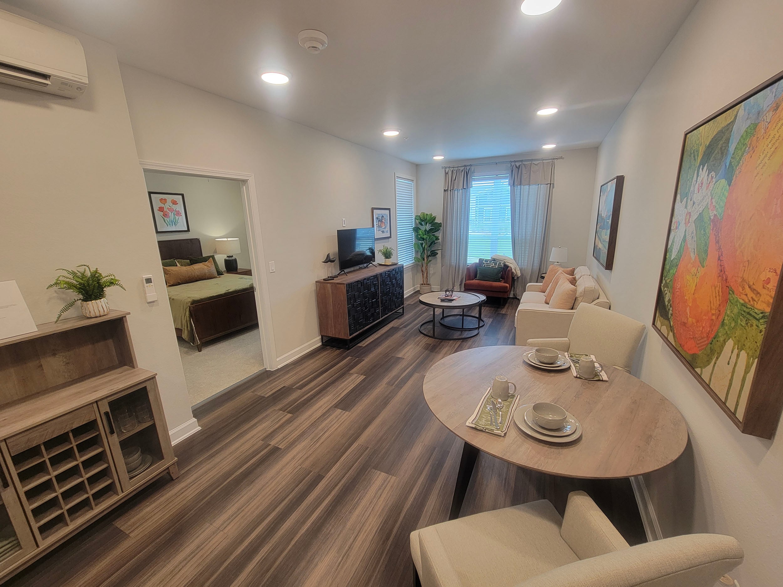 Modern living room and dining area featuring a cozy sofa, stylish coffee table, and dining set. The space includes a view into a bedroom, decorative artwork, and an entertainment center with a television. Natural light streams through the window, enhancing the inviting atmosphere.