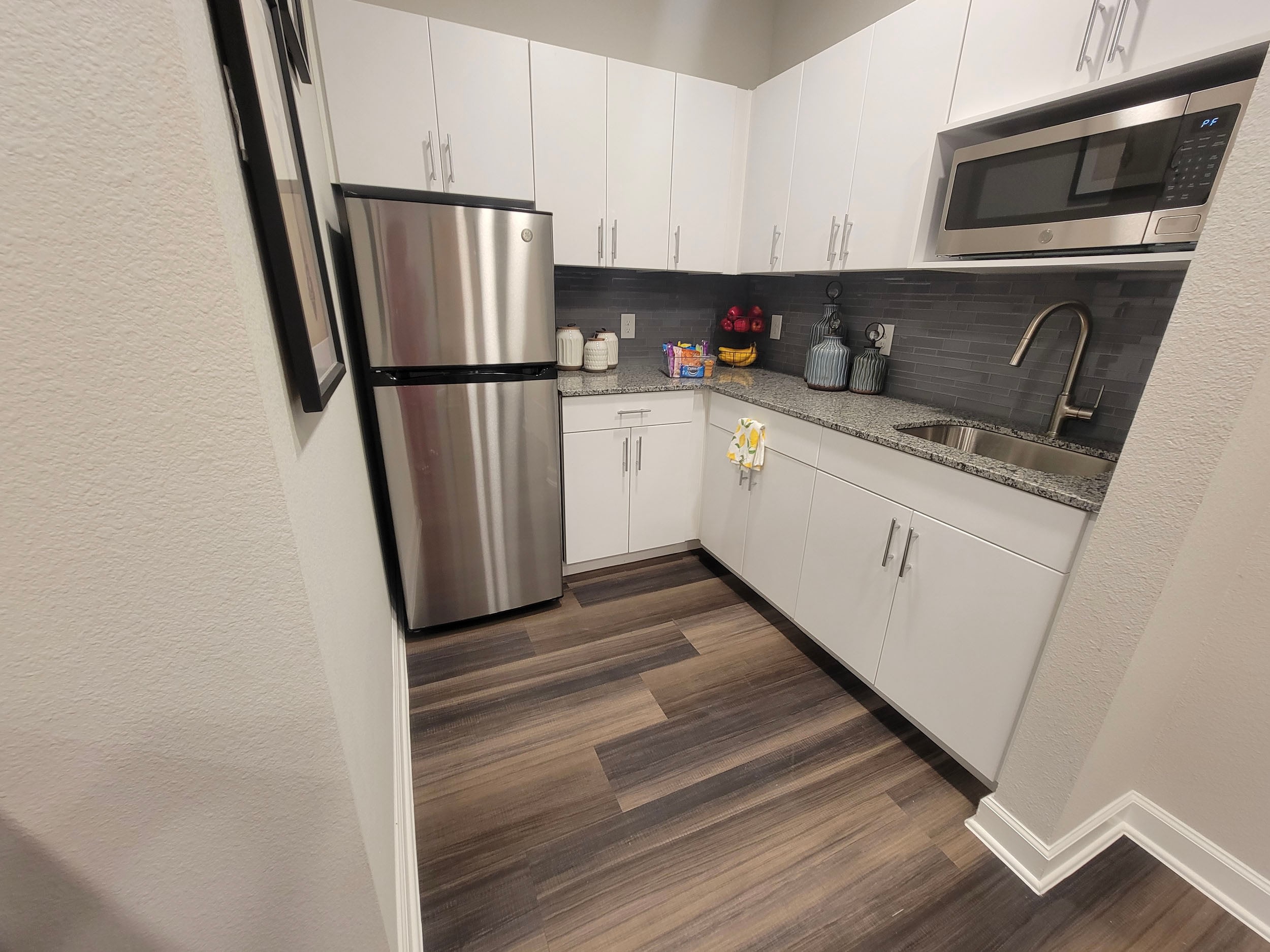 Modern kitchen featuring stainless steel refrigerator, microwave, and sleek white cabinetry with granite countertops. The space includes decorative items and a stylish backsplash, complemented by warm wood-like flooring. Ideal for contemporary living.