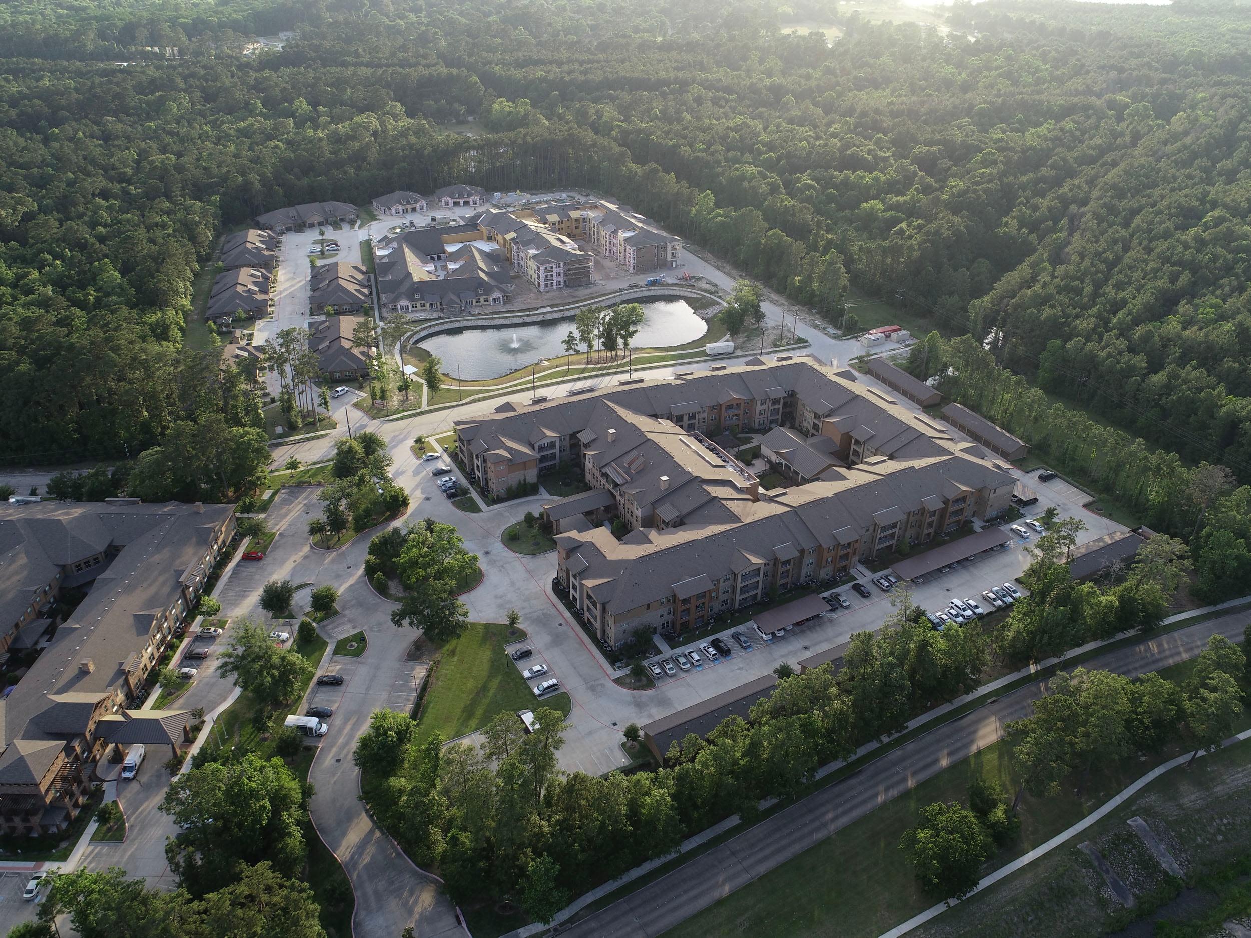 Aerial view of a modern residential complex surrounded by lush greenery, featuring multiple buildings, a pond with a fountain, and landscaped pathways, highlighting a peaceful community setting.