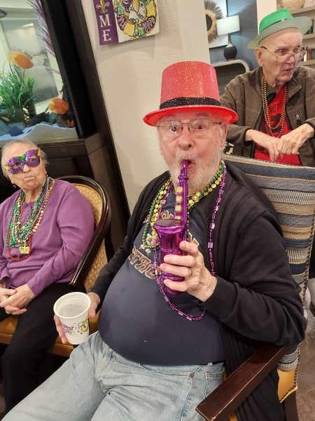 Elderly man in festive hat and Mardi Gras beads enjoying a celebration activity at senior living facility