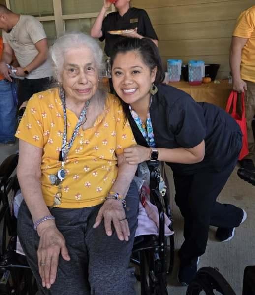 Elderly woman in yellow shirt sitting with younger female caregiver in dark clothing, both smiling