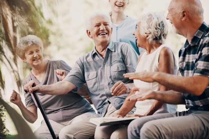 Group of seniors sitting together outdoors, laughing and socializing with a caregiver standing behind them