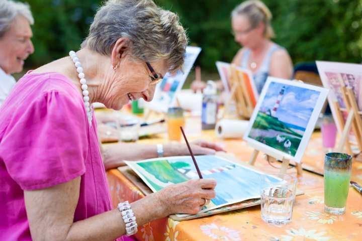 Senior woman in pink shirt painting on canvas at an outdoor art class with other seniors in the background
