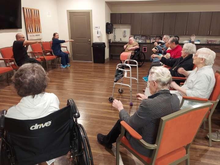 Group of seniors sitting in a circle in a common area with walkers and wheelchairs, participating in an activity