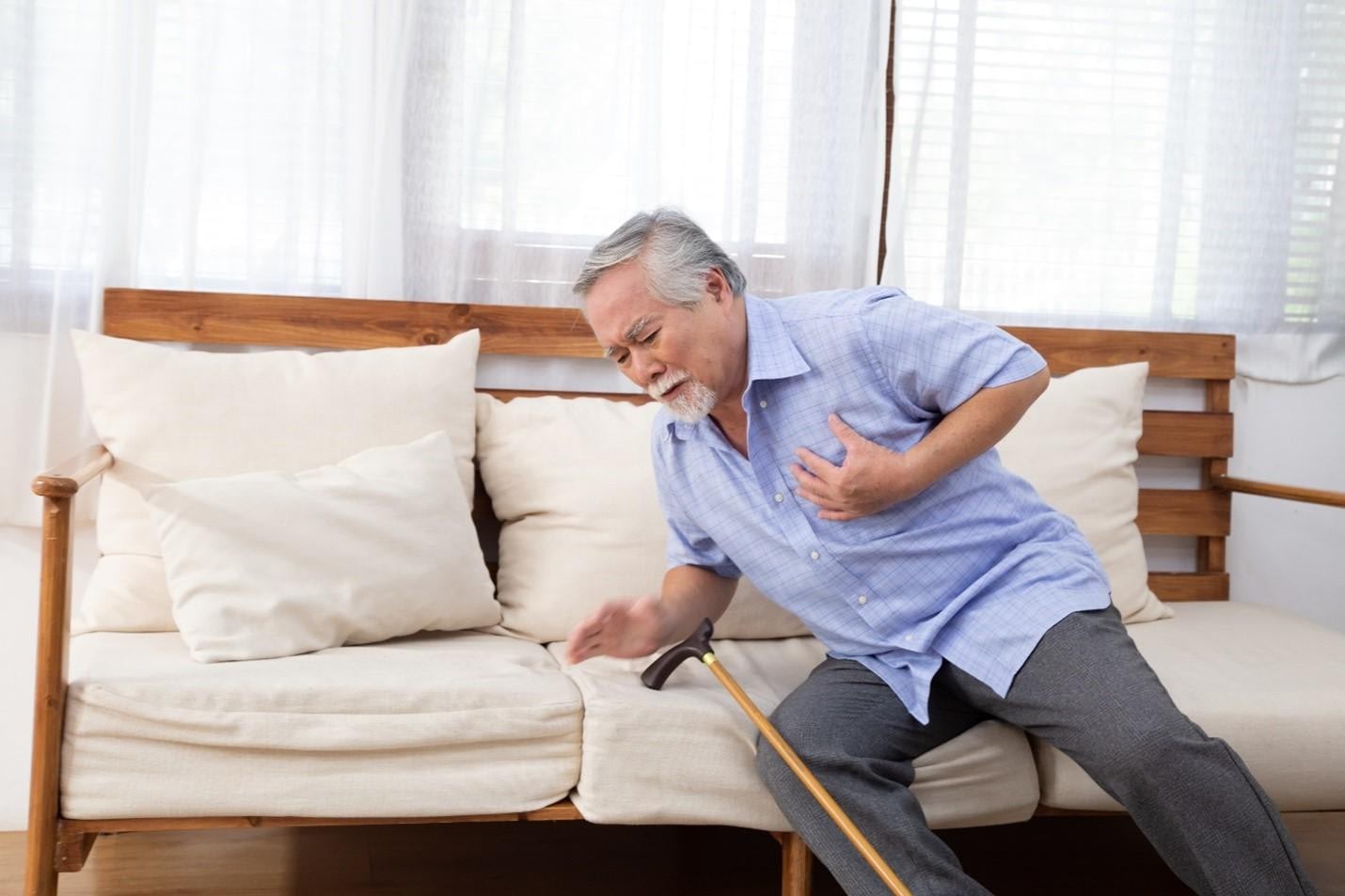 An elderly man holding his chest while sitting on a chair, appearing to be in distress or experiencing pain